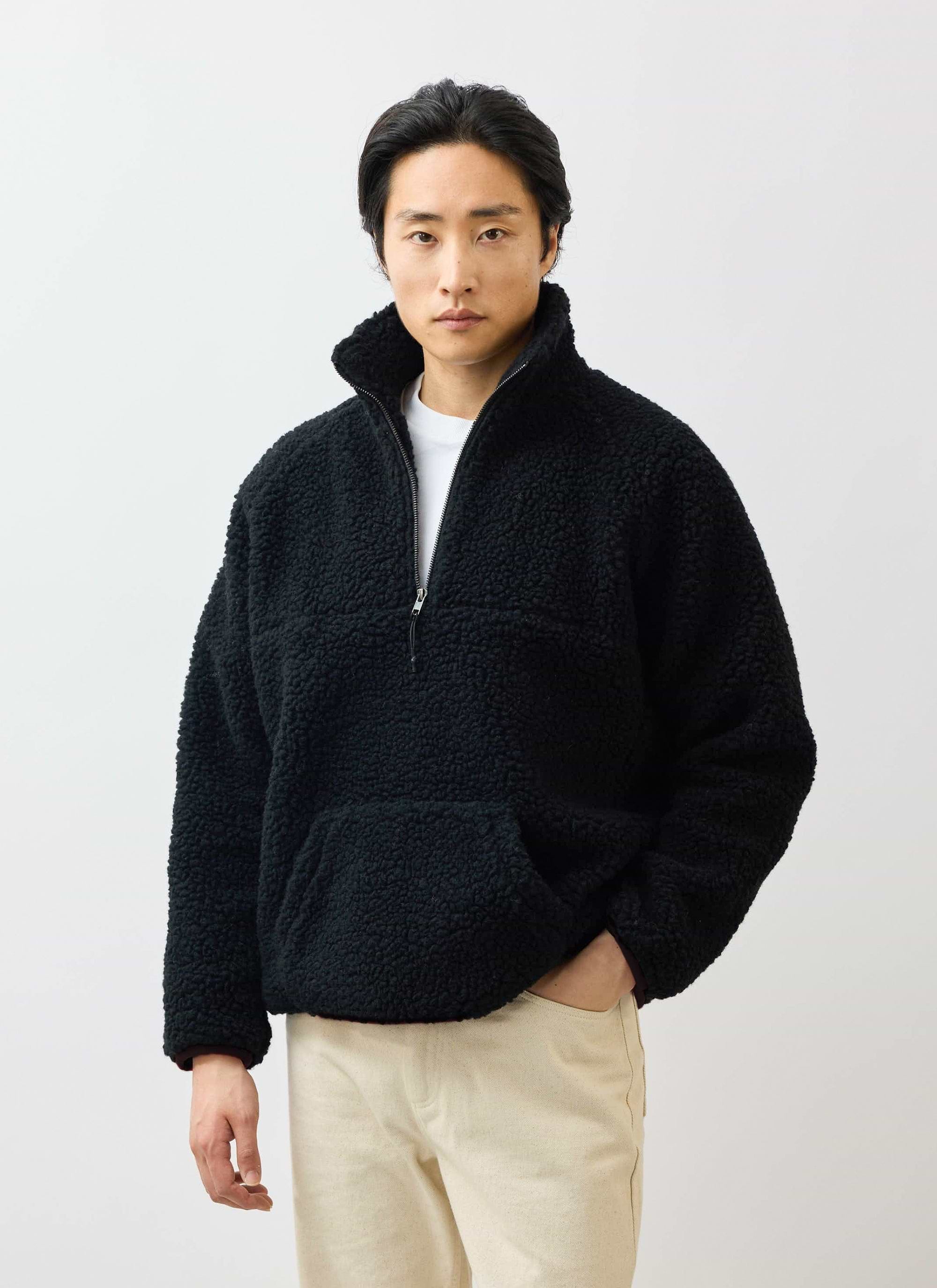 A model wears the Percival Quarter Zip Wool Fleece Pullover in black, styled with a white t-shirt and cream trousers.