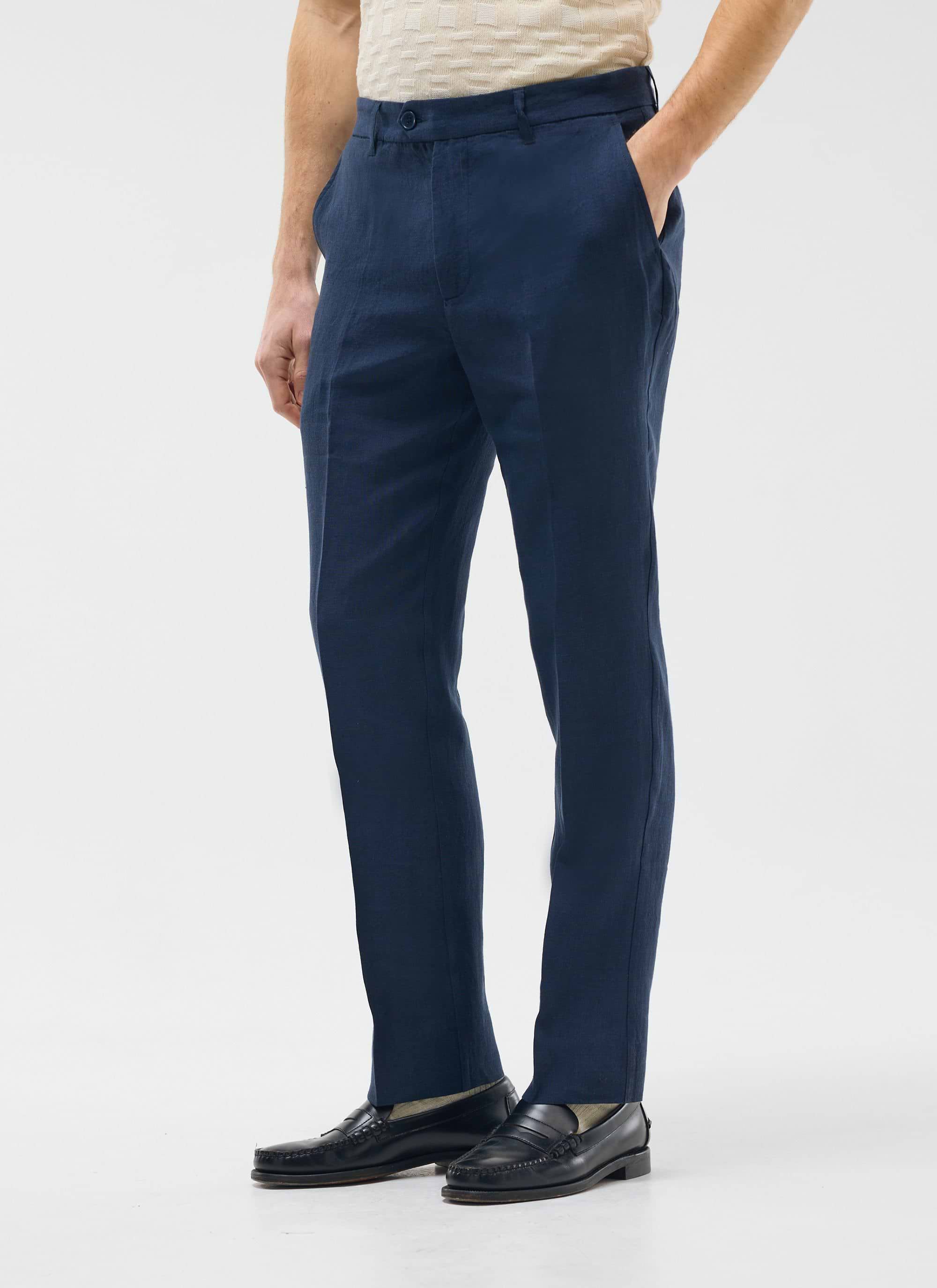 Model wears Percival navy tailored linen trousers with a cream top and black loafers on a white background.