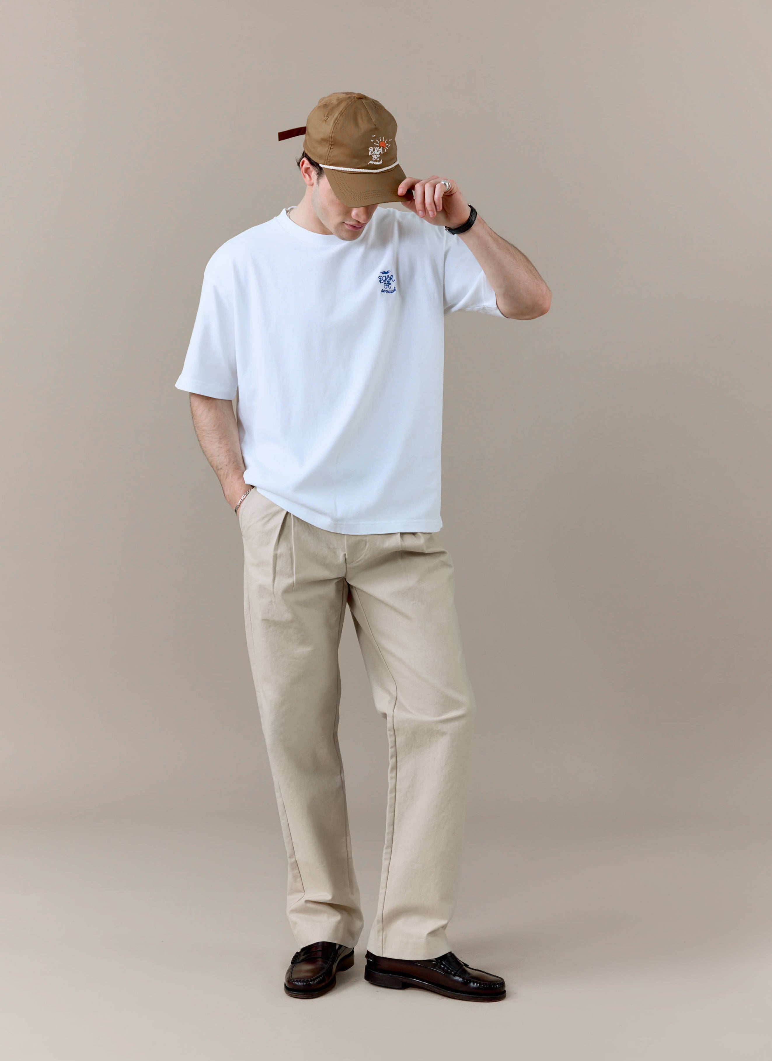 A model styles the white Percival x Brighton & Hove Albion F.C. Archive Kits T-Shirt with beige trousers and a cap.