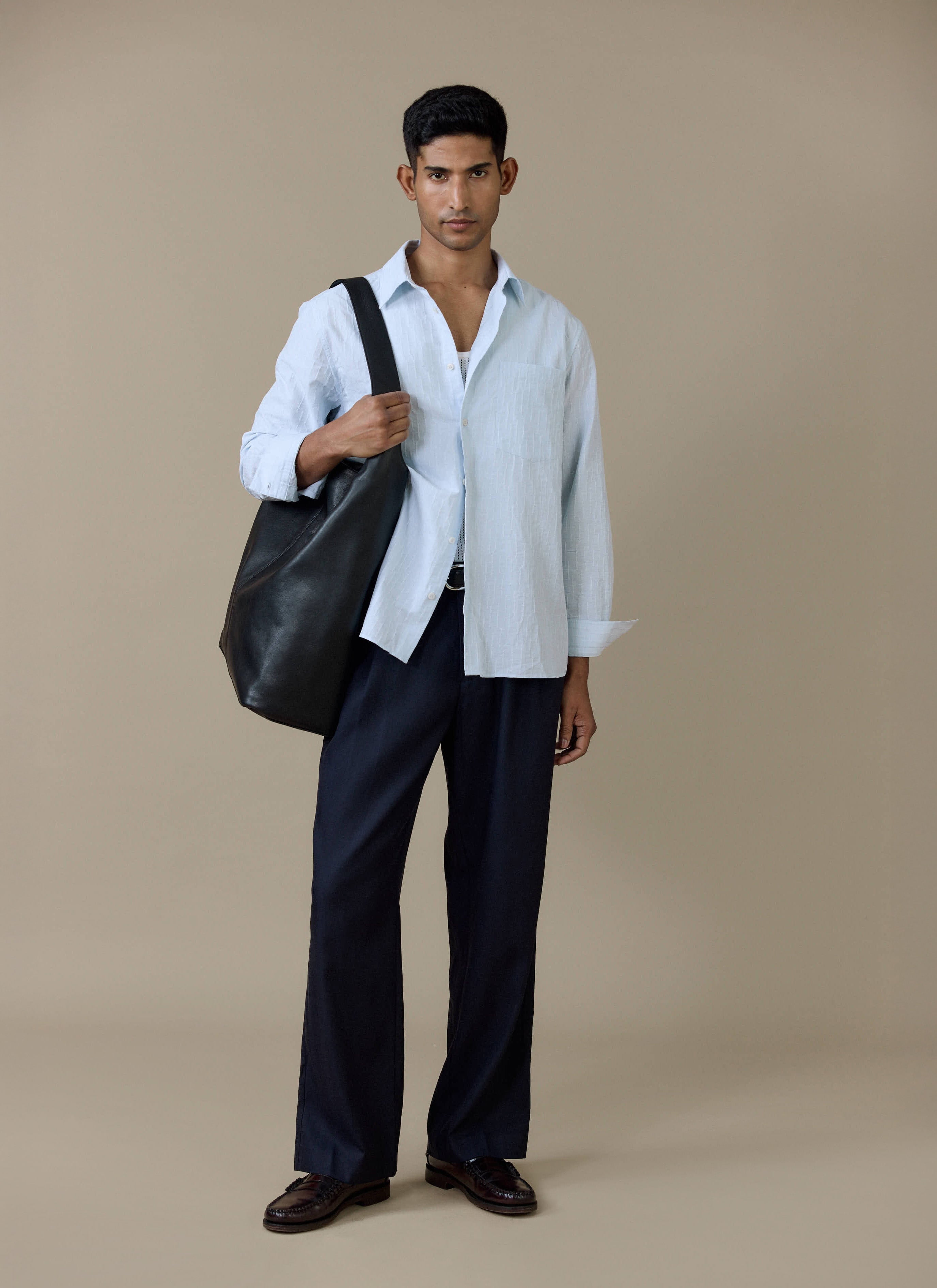 A model wearing the light blue Percival Paros Pearce Shirt in cotton seersucker with navy trousers and a black leather bag.