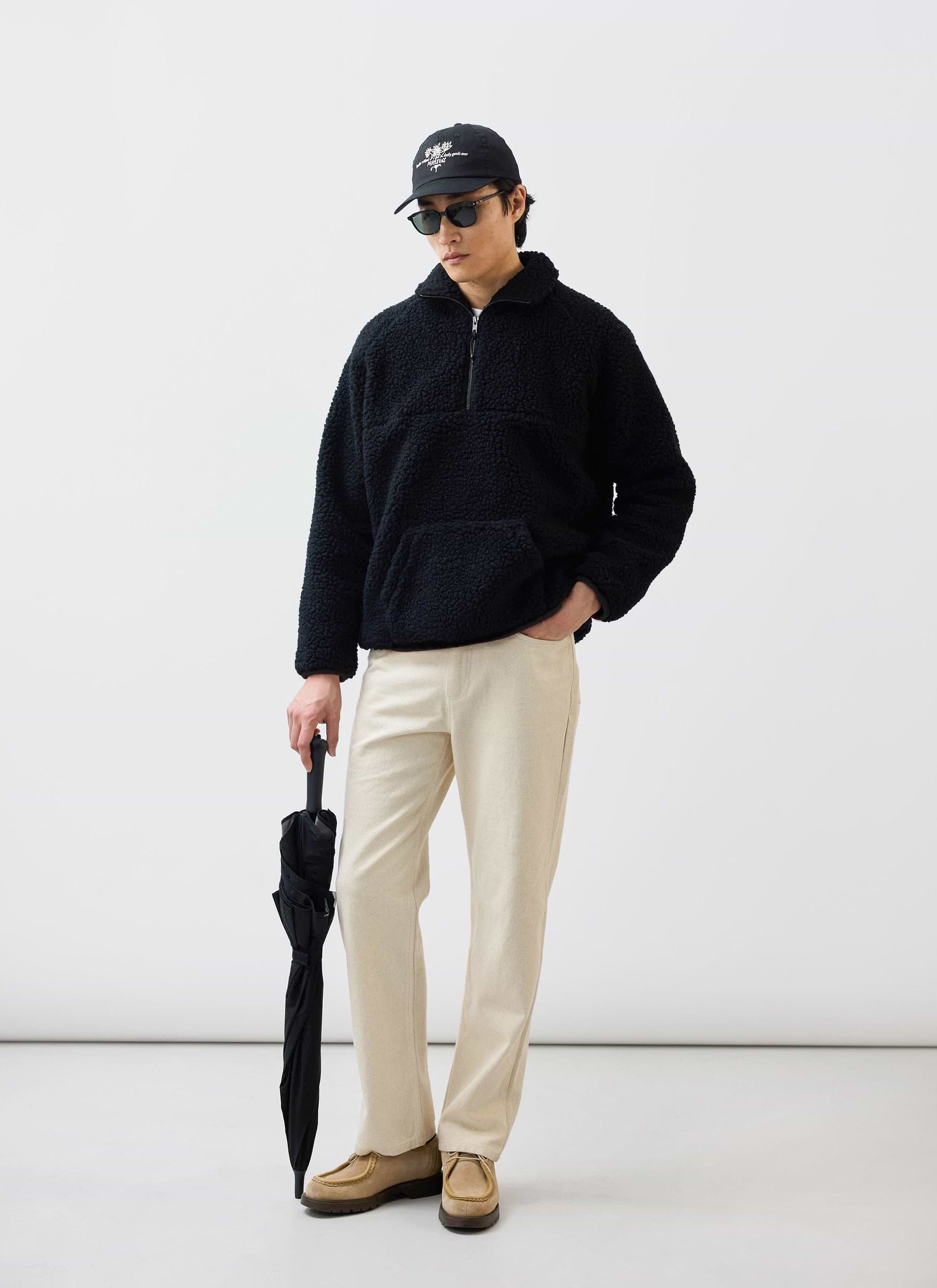 A man models the Percival black Quarter Zip Wool Fleece, styled with cream trousers, a cap and holding an umbrella.
