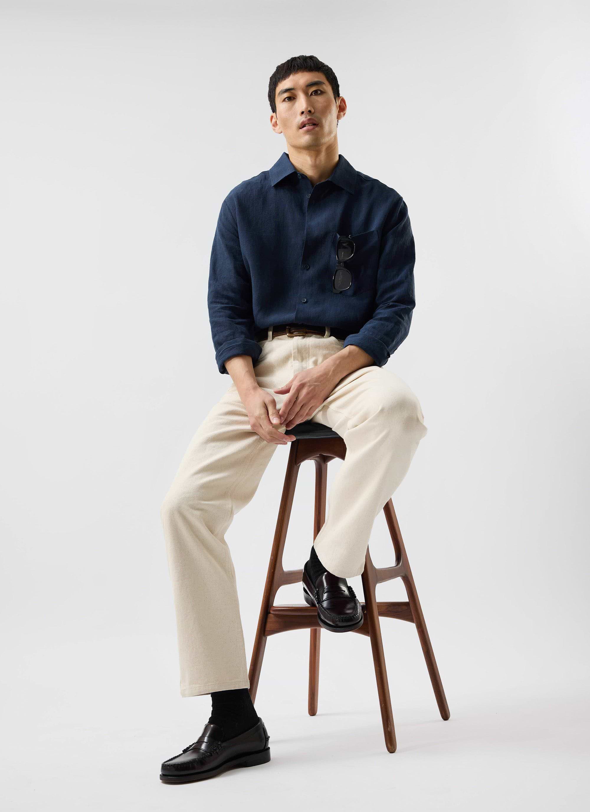 A model sits on a wooden stool wearing the navy linen Percival Pearce Shirt, paired with cream trousers and loafers.