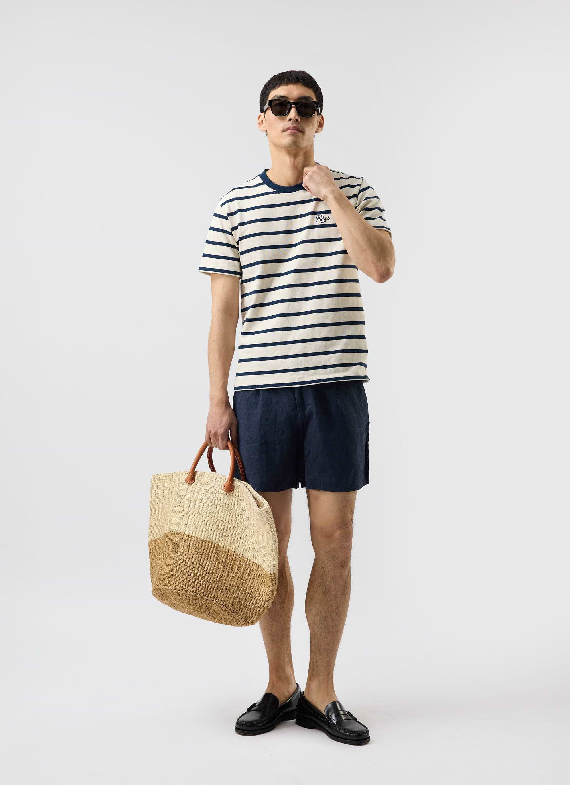 A model wears the Percival Breton Stripe T Shirt in ecru and navy, styled with navy shorts, sunglasses, and a woven tote bag.