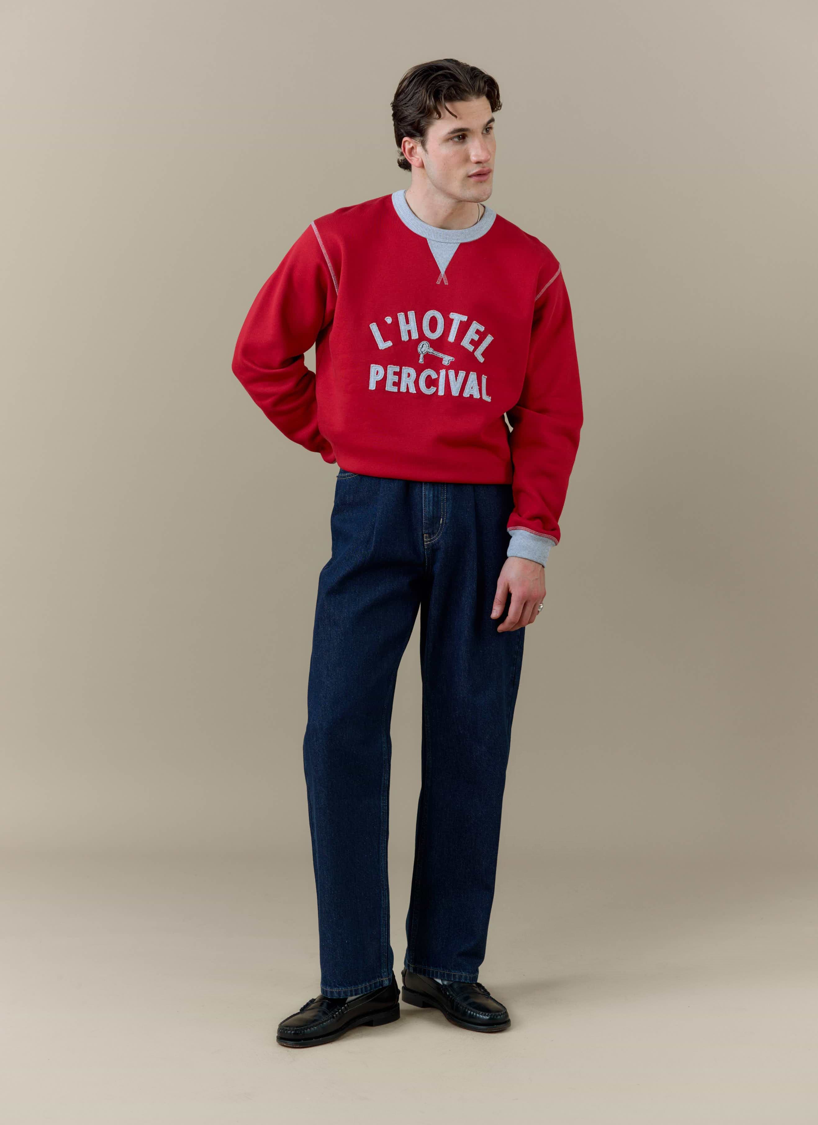 A model wearing the red Ronald sweatshirt from Percival with 'L'Hotel Percival' applique and dark blue trousers.