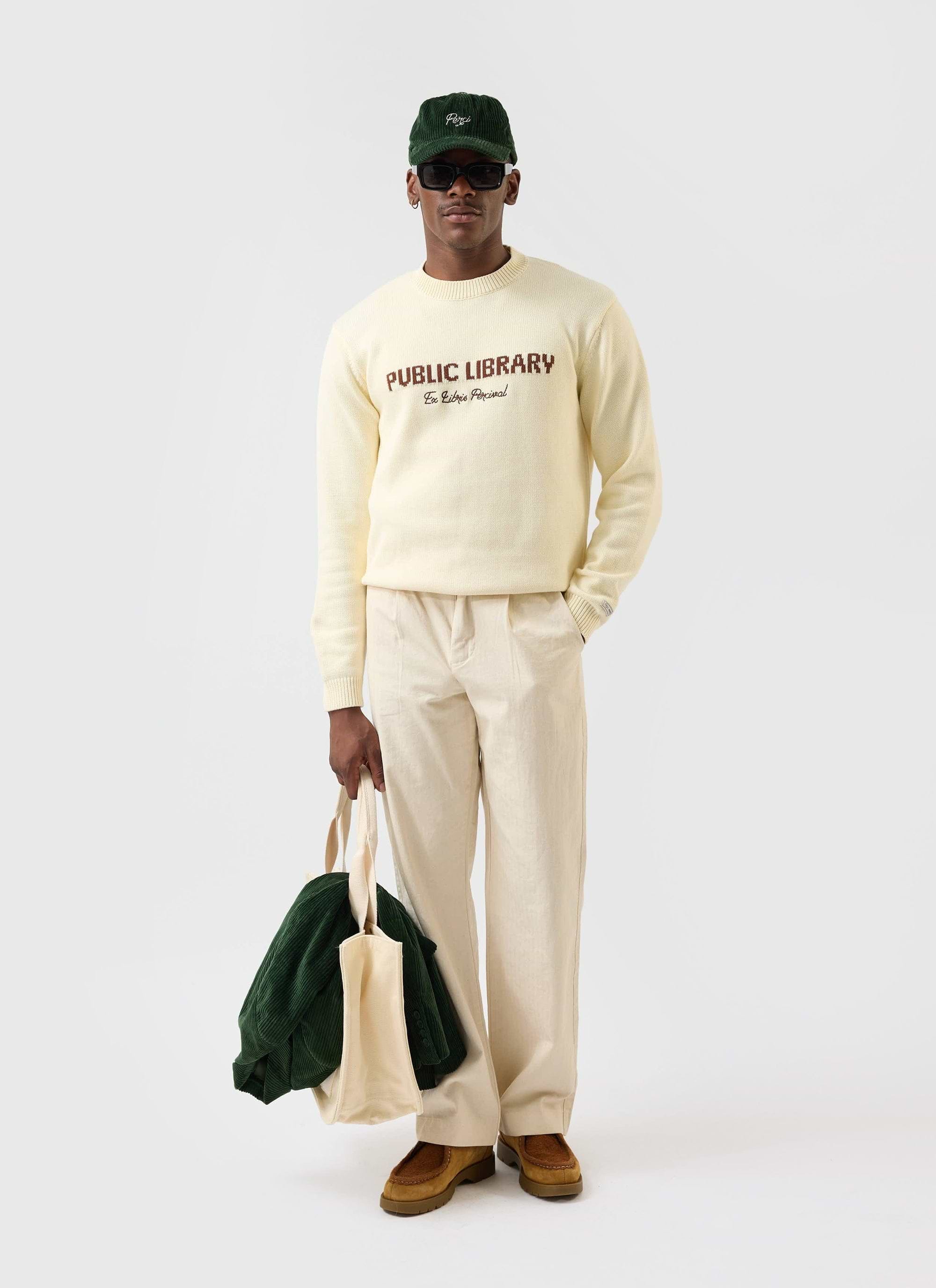 A model wears the ecru cream Public Library knitted jumper from Percival, paired with cream trousers and a green cord cap.