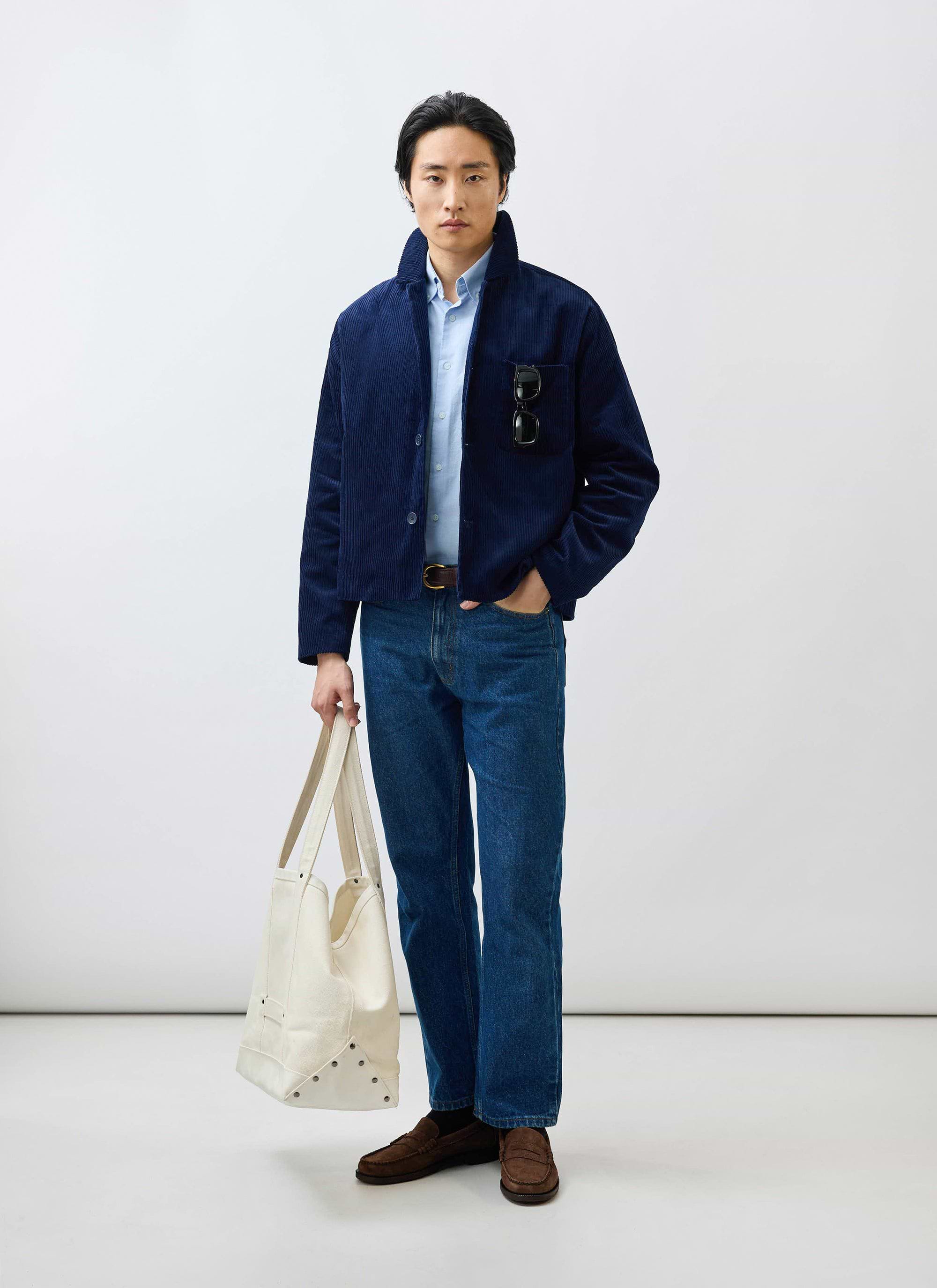 A male model wears the Percival navy corduroy overshirt layered over a blue shirt, with blue jeans and a cream tote bag.