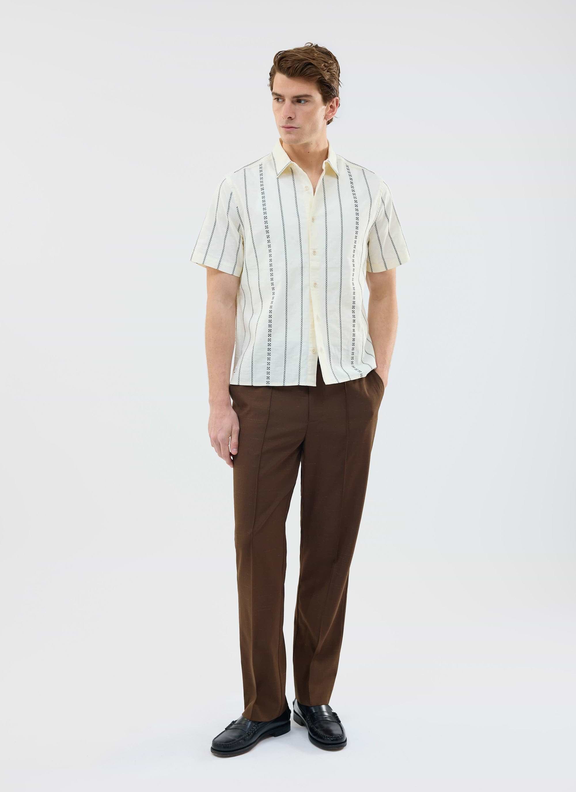 A model wears the ecru Cross Stitch Boxy Clerk Shirt from Percival, styled with smart brown trousers and black loafers.