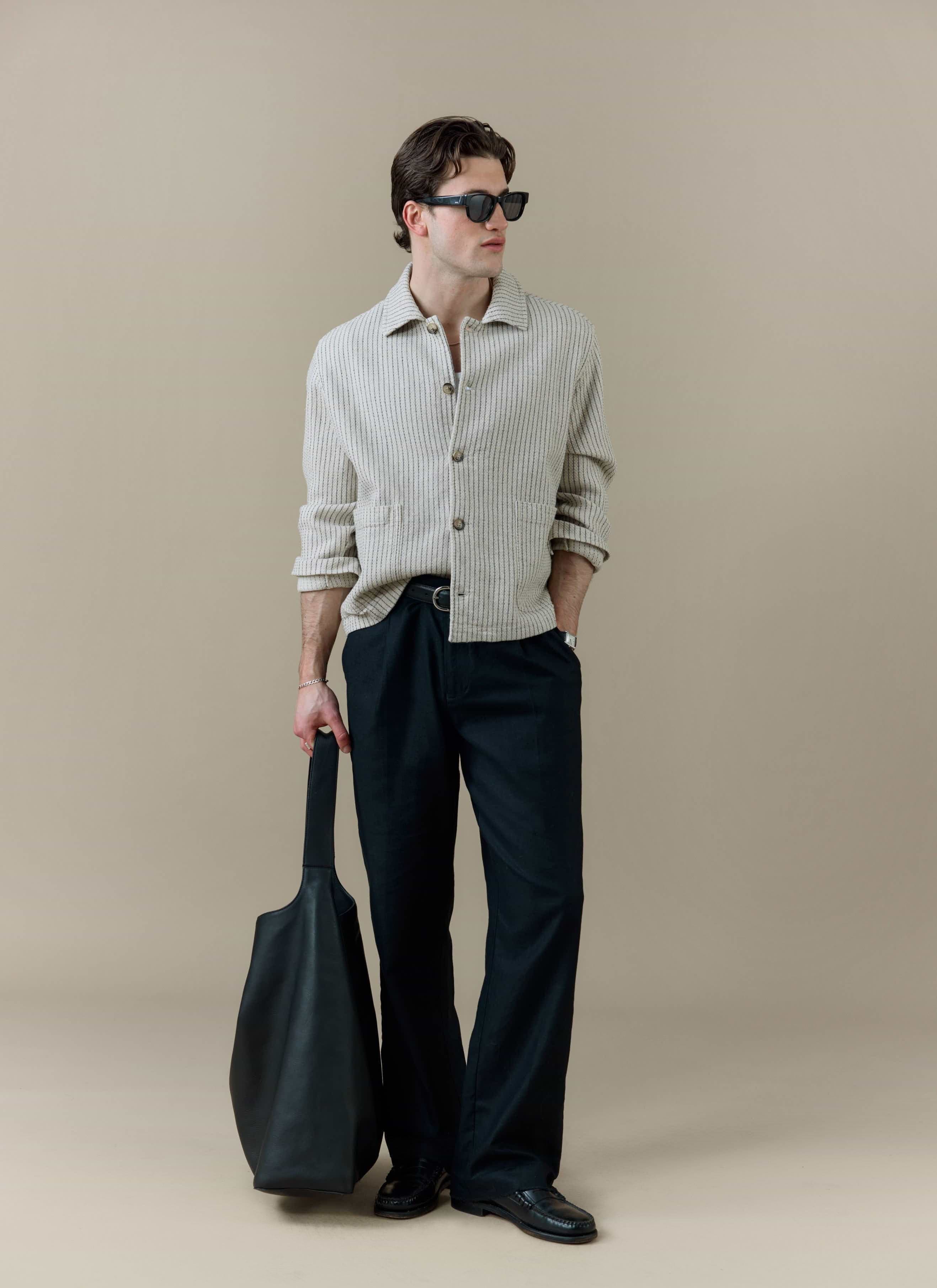 Model wearing the Percival Sorrento Boxy Overshirt in cream linen, paired with black trousers and a large black bag.