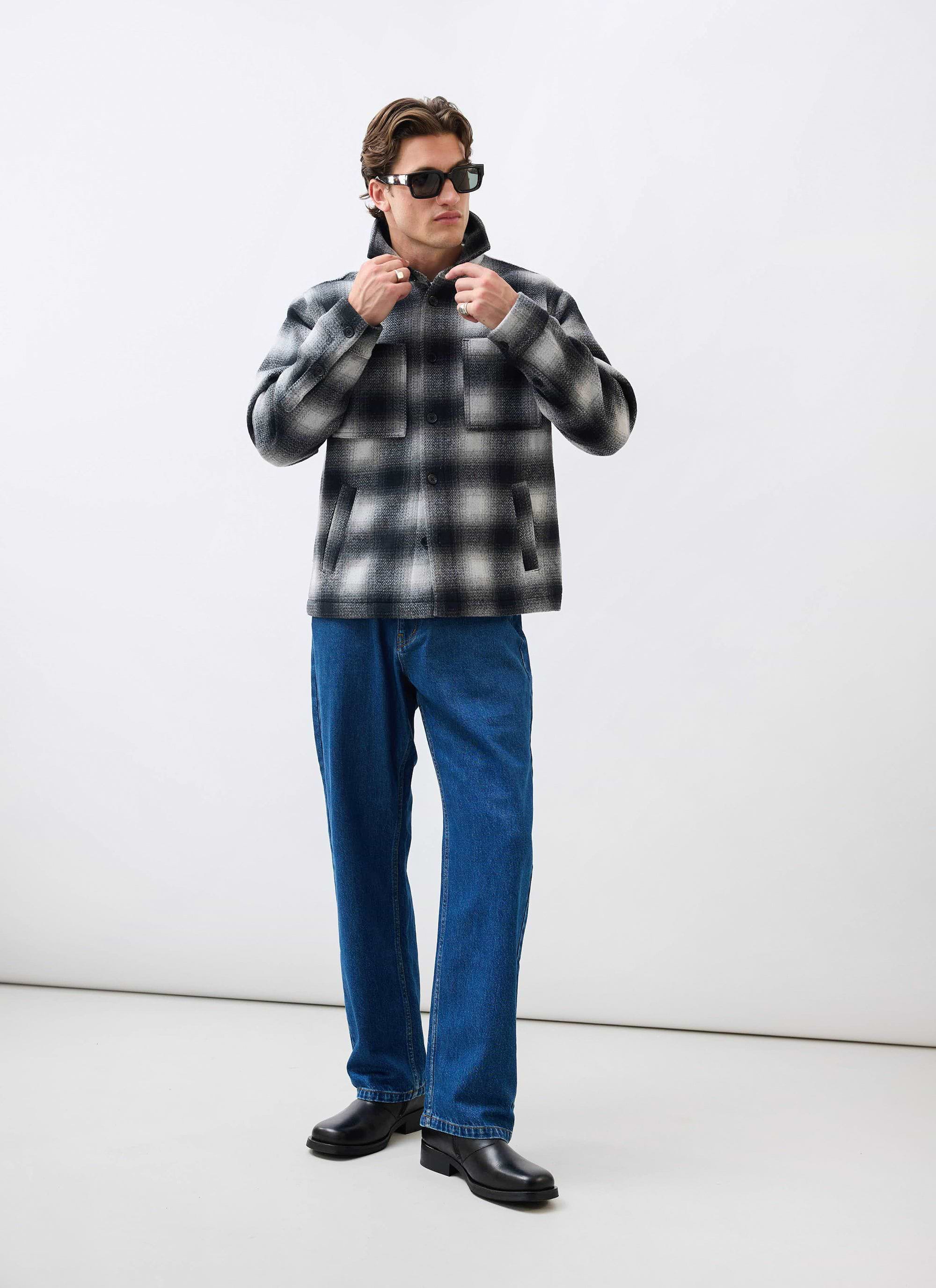 A man models the Percival Check Overshirt in grey, styled with dark blue jeans, black shoes, and sunglasses.