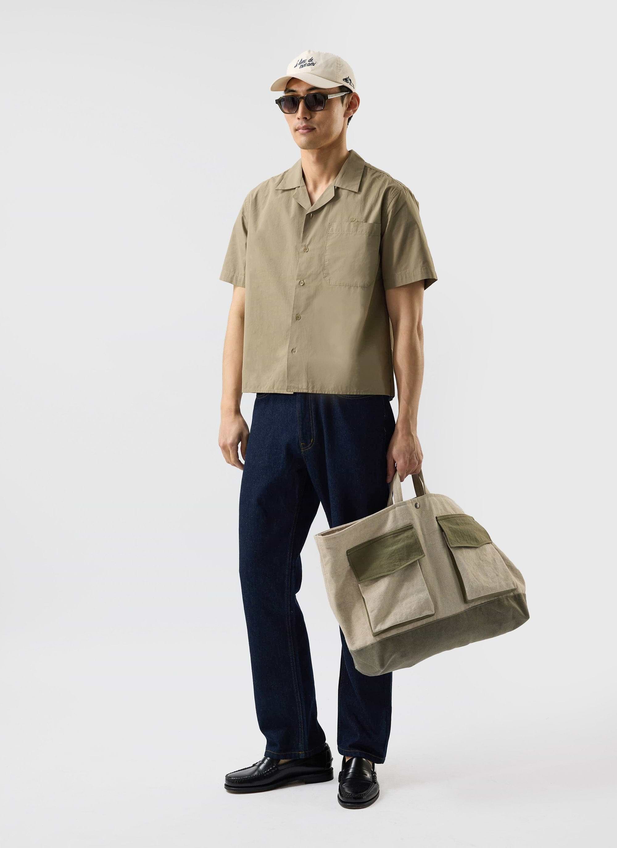 A man models the olive Boxy Cropped Cuban Collar Shirt from Percival with dark denim jeans, sunglasses, and a cap.