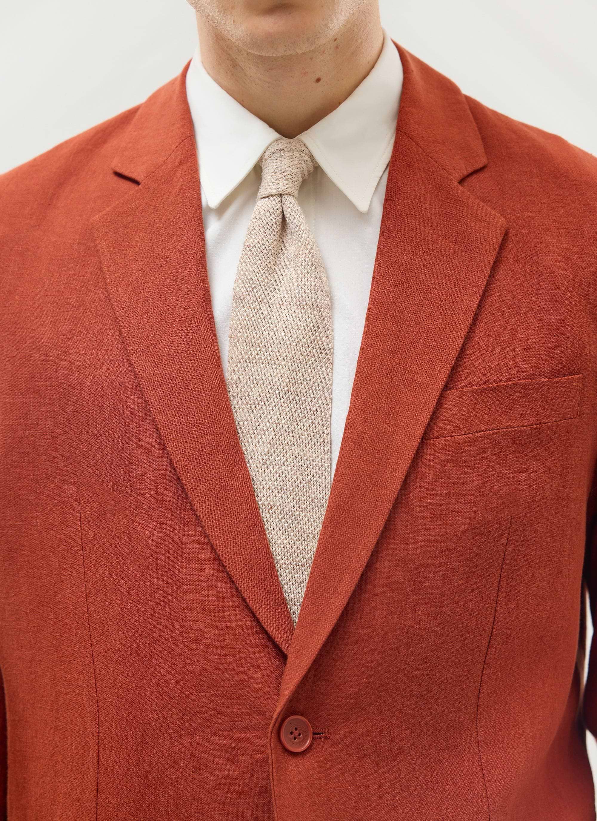 Close up of the Percival Relaxed Tailored Linen Blazer in rust, worn over a white shirt and beige knitted tie.