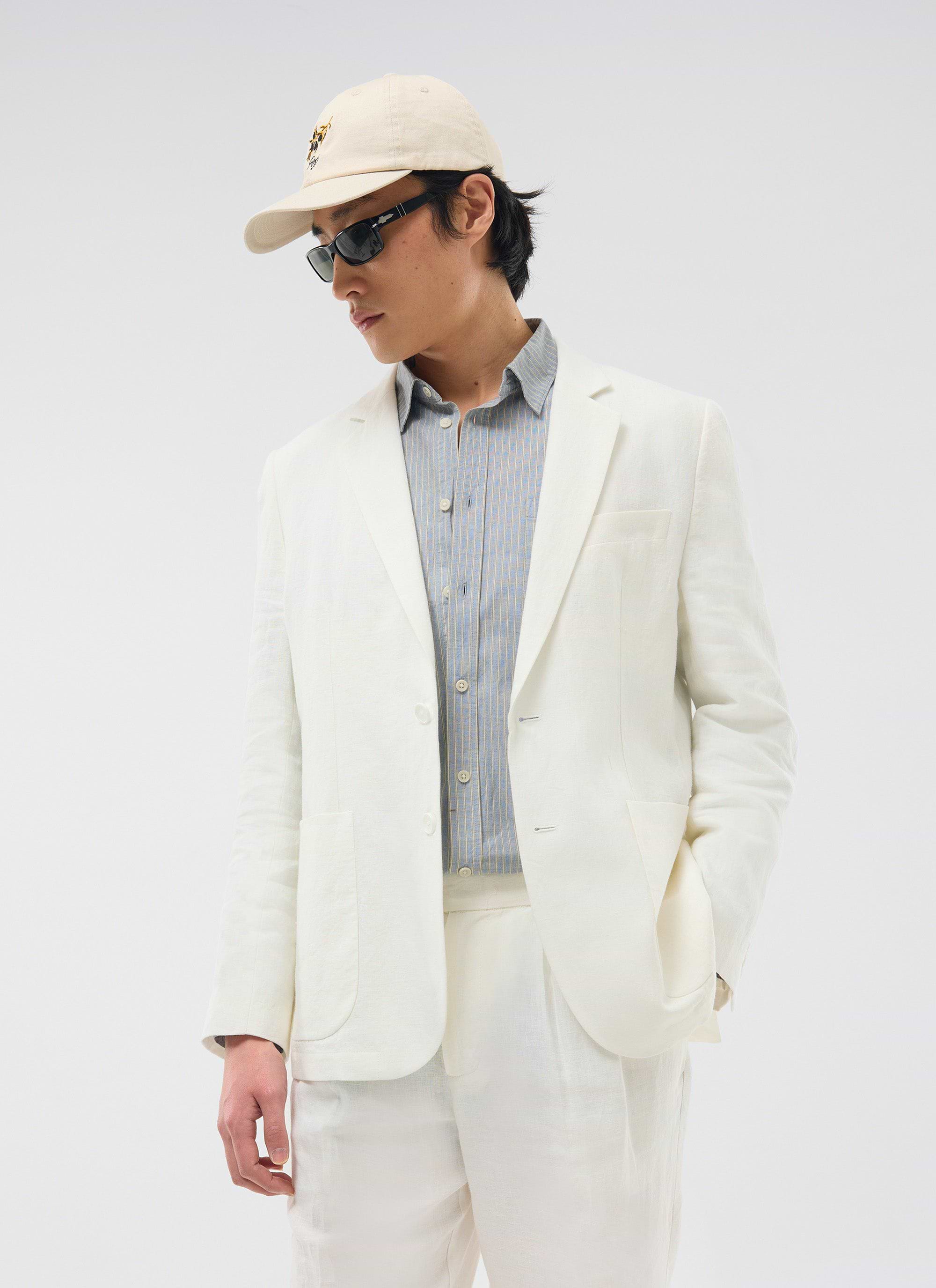 A model wears the Percival Relaxed Tailored Linen Blazer in white, styled casually with a striped shirt and a baseball cap.