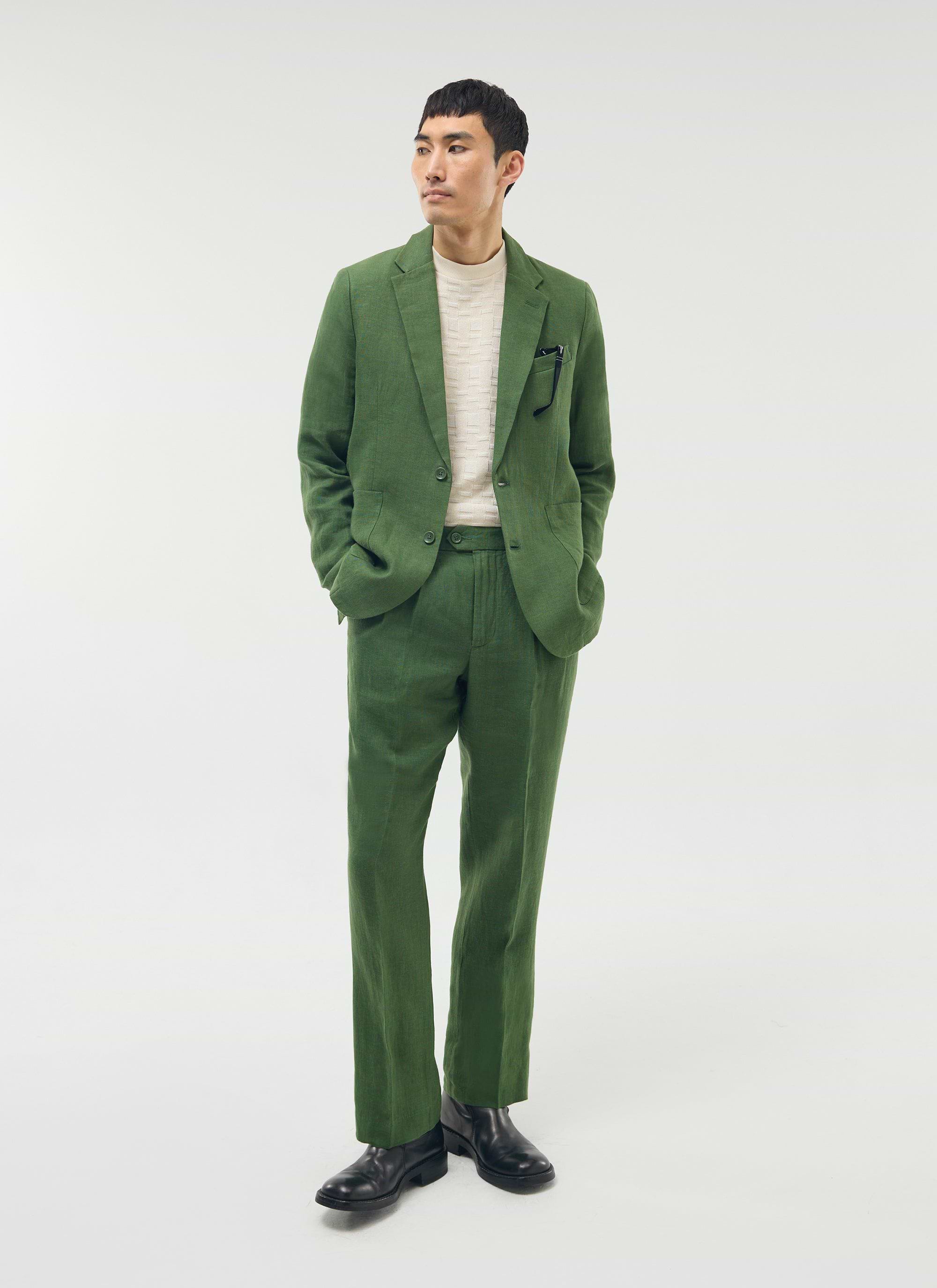 A model wears Percival's forest green linen pleated trousers with the matching blazer, a cream knit and black boots.