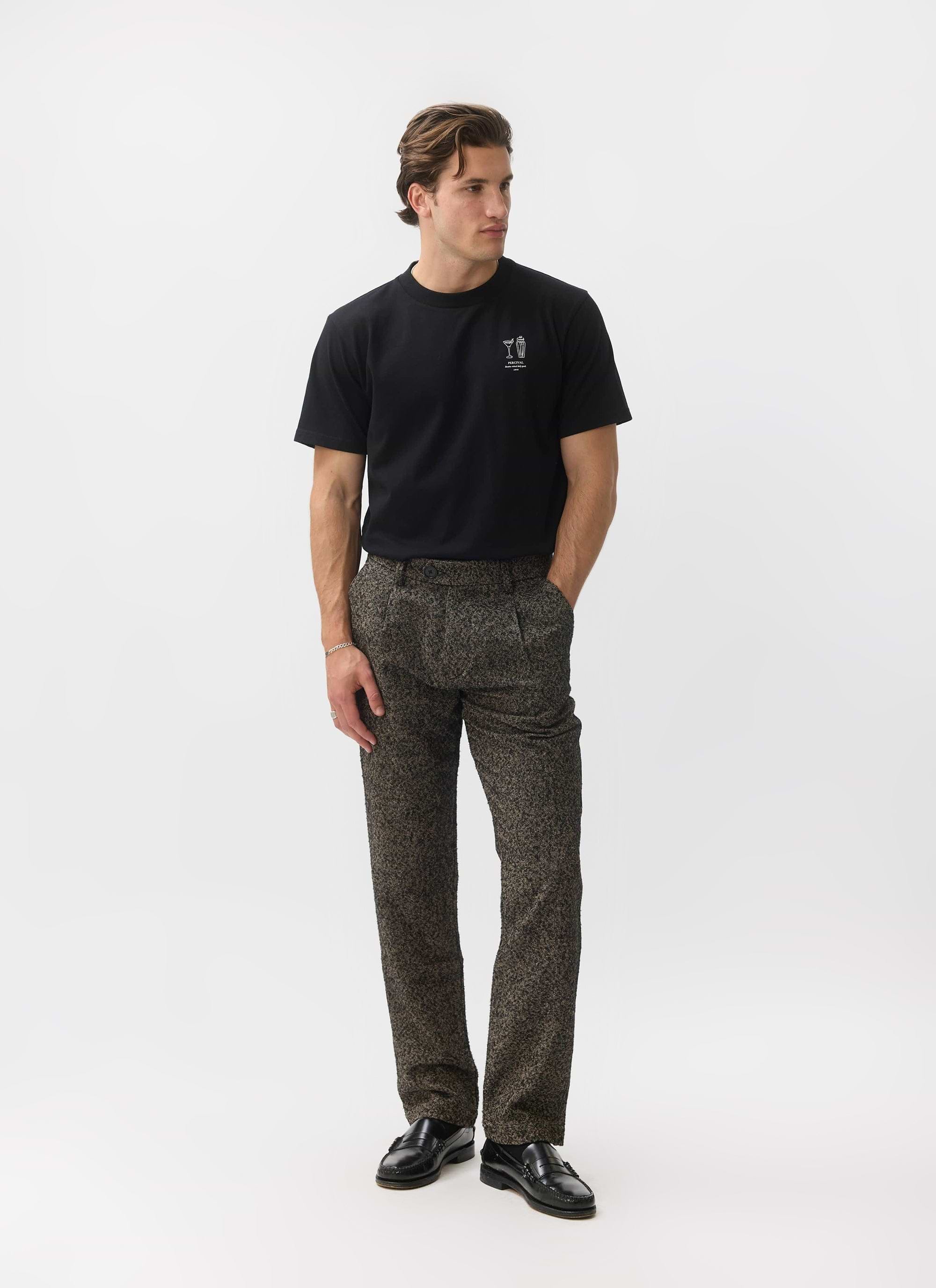A man models the Percival Blyton Trousers in a warm grey nep fabric against a white studio background.