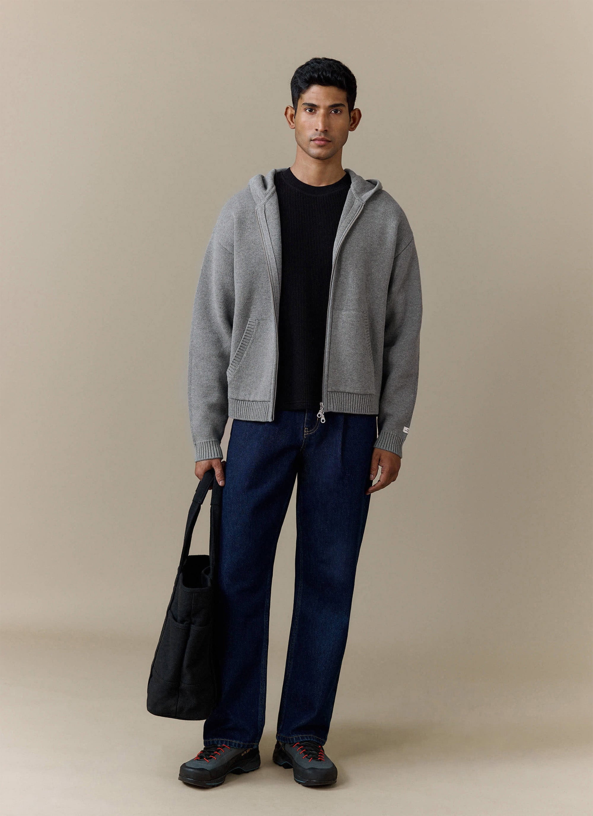 Full shot of a model wearing the Percival Chronicle Knitted Hoodie in grey, layered over a black tee with dark denim jeans.