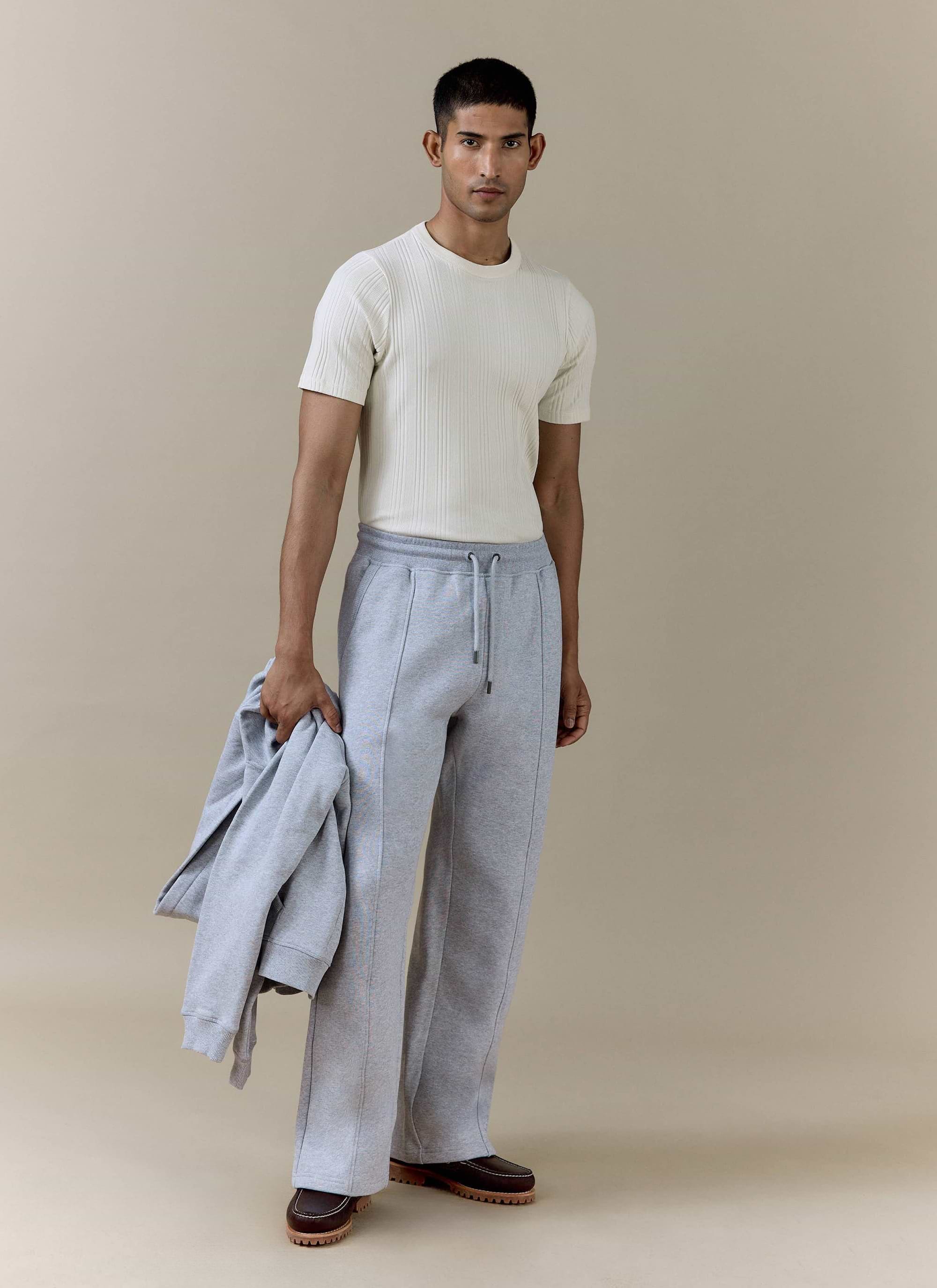 Model wears the Percival cream Leon Ribbed T Shirt and grey cotton Chronicle Straight Leg Trackpants in a studio.