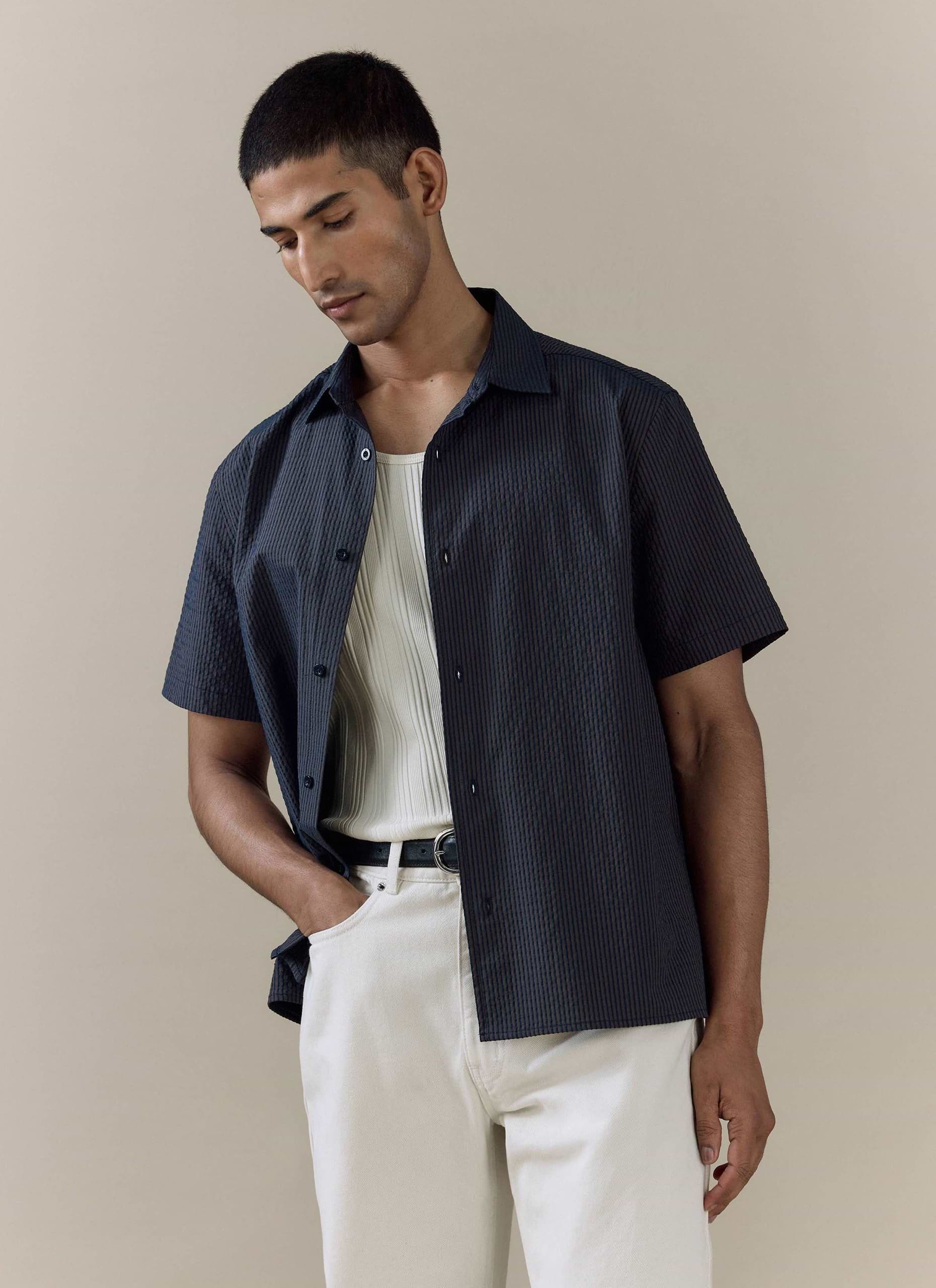 Model wears the navy stripe Rowan Seersucker Boxy Shirt by Percival, styled with a white ribbed vest and trousers.