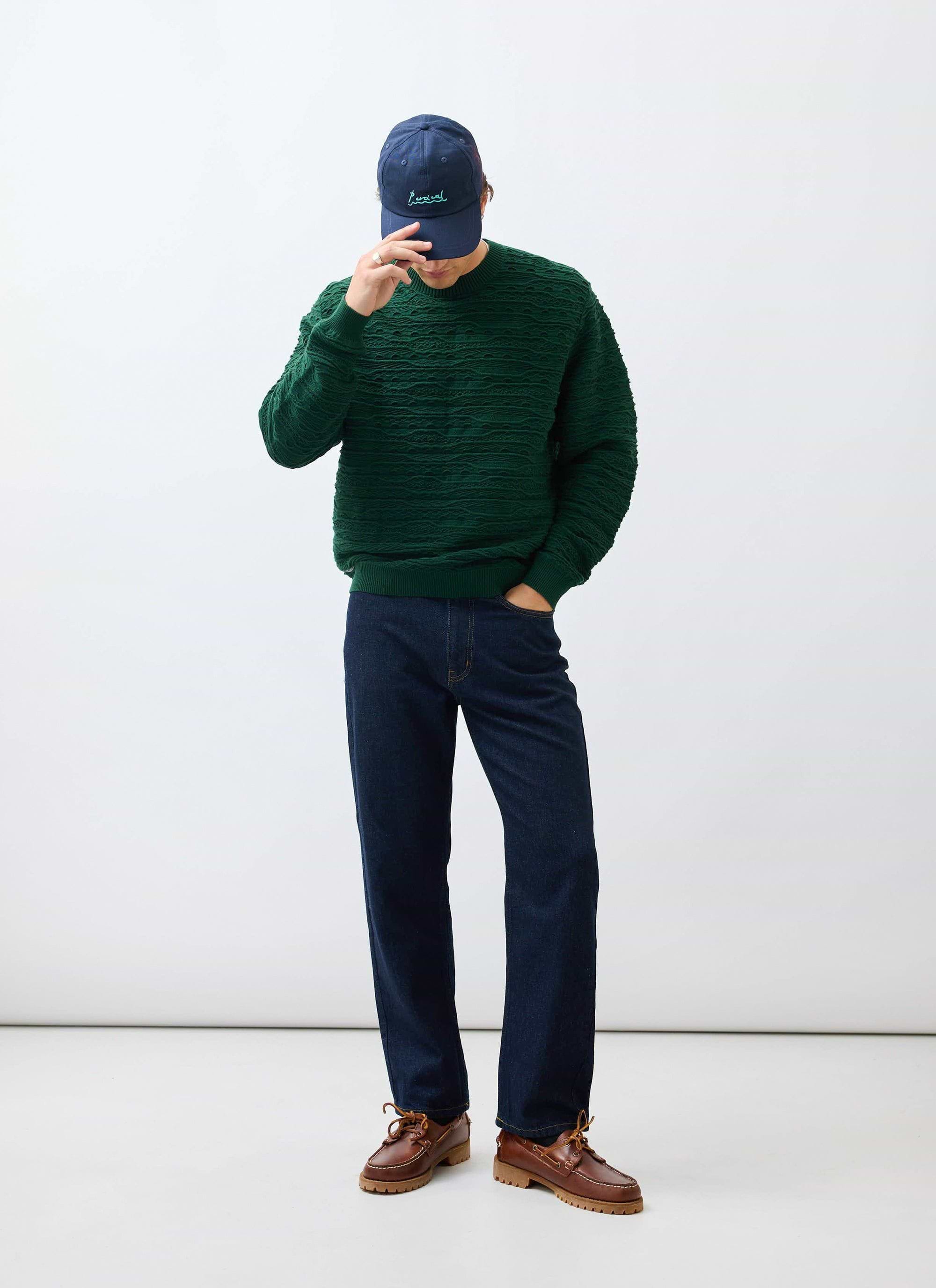 A model wears the Percival Tales Knitted Crew Neck Jumper in forest green, styled with dark denim and a navy Percival cap.
