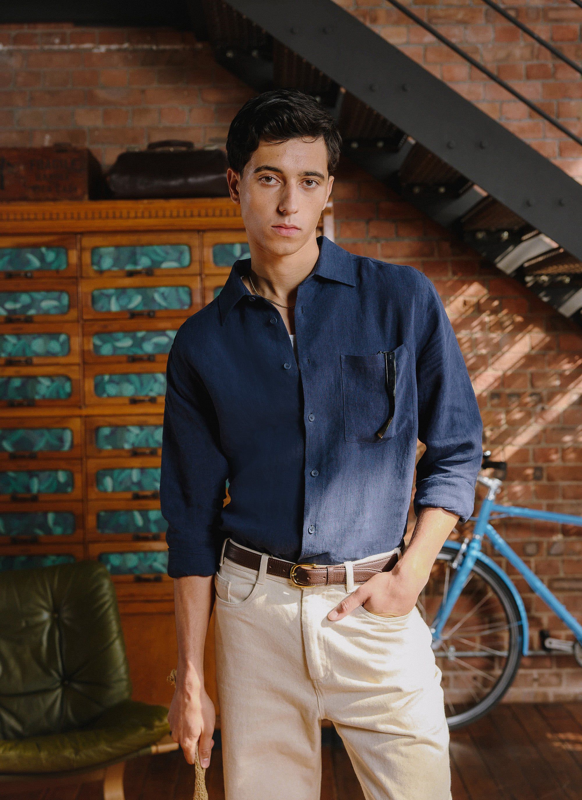 A man models the Percival Pearce Shirt in navy linen with cream trousers in a sunlit room with exposed brick walls.