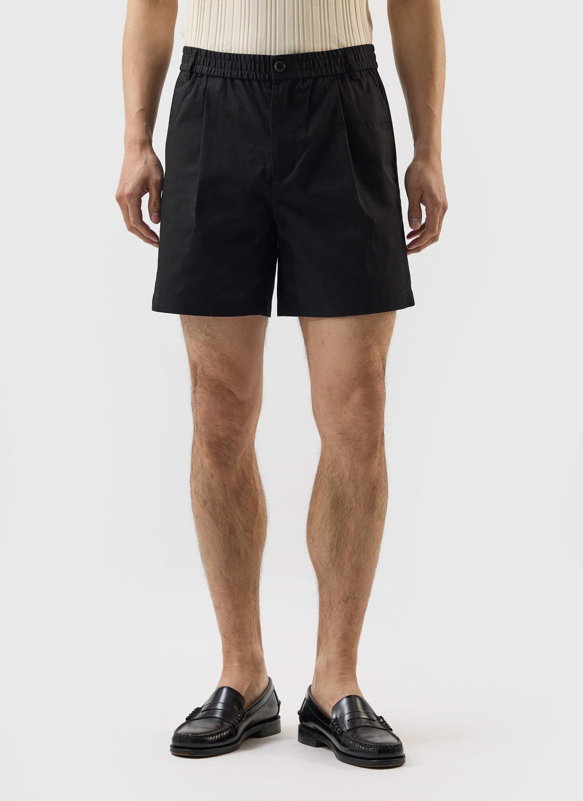 A model wears the Percival Enzo Tailored Shorts in black cotton with a cream ribbed top and black leather loafers.
