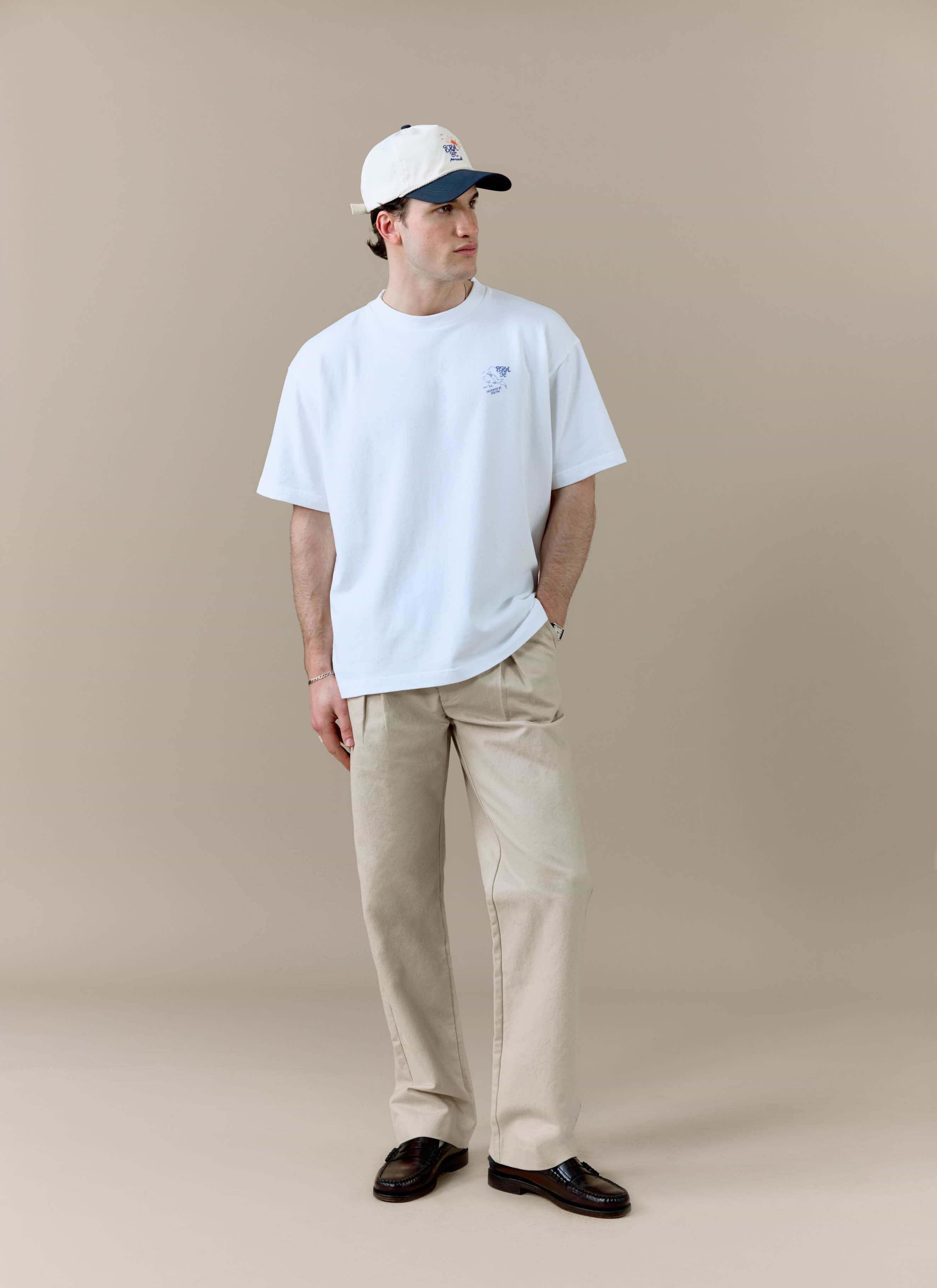 A model styled in the white Percival x Brighton & Hove Albion F.C. Beach Front T-shirt with beige trousers and a cap.