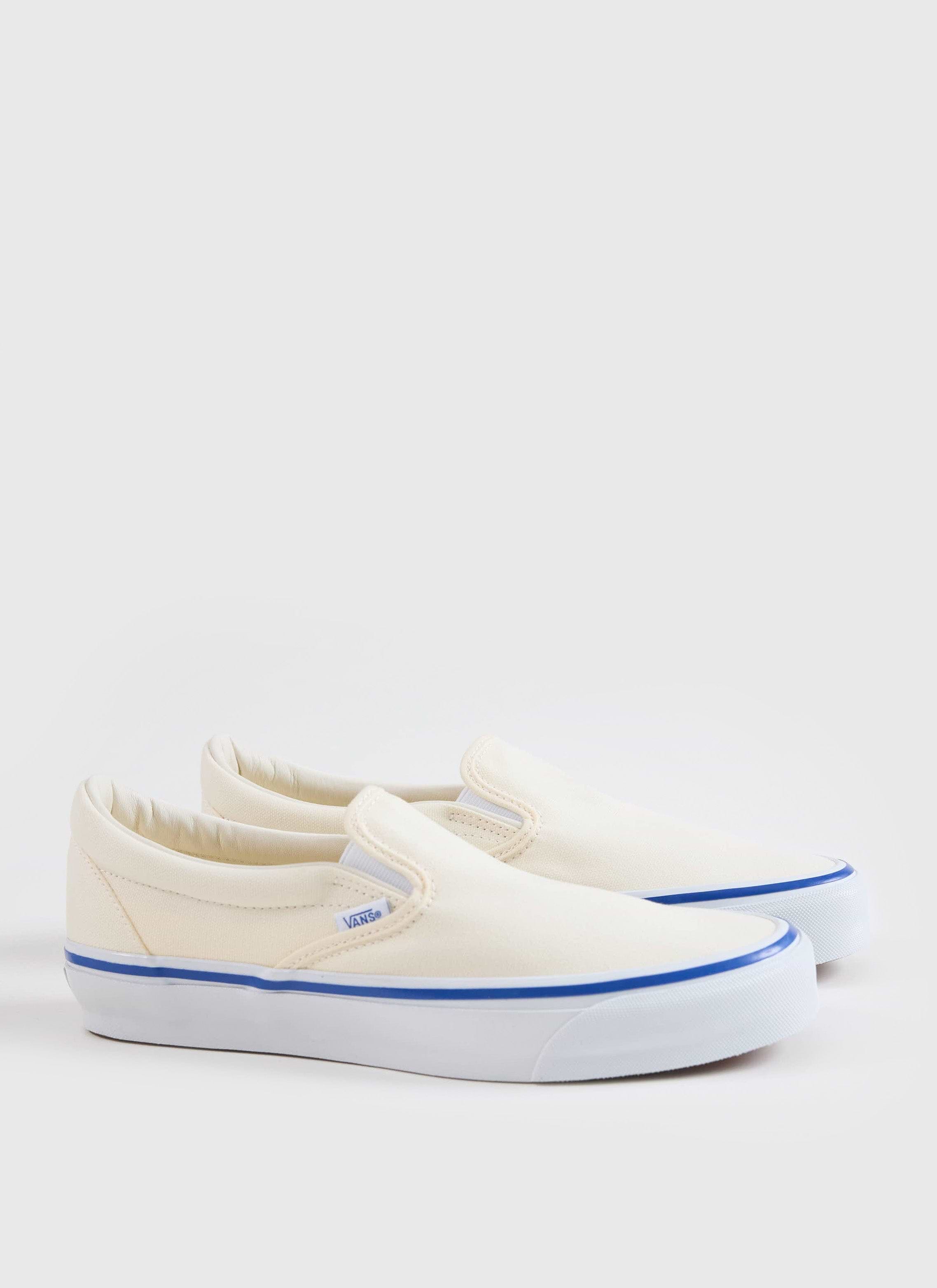 A side view of the Vans Slip-On Reissue 98 Shoes in off white canvas with a blue contrast stripe, from Percival.