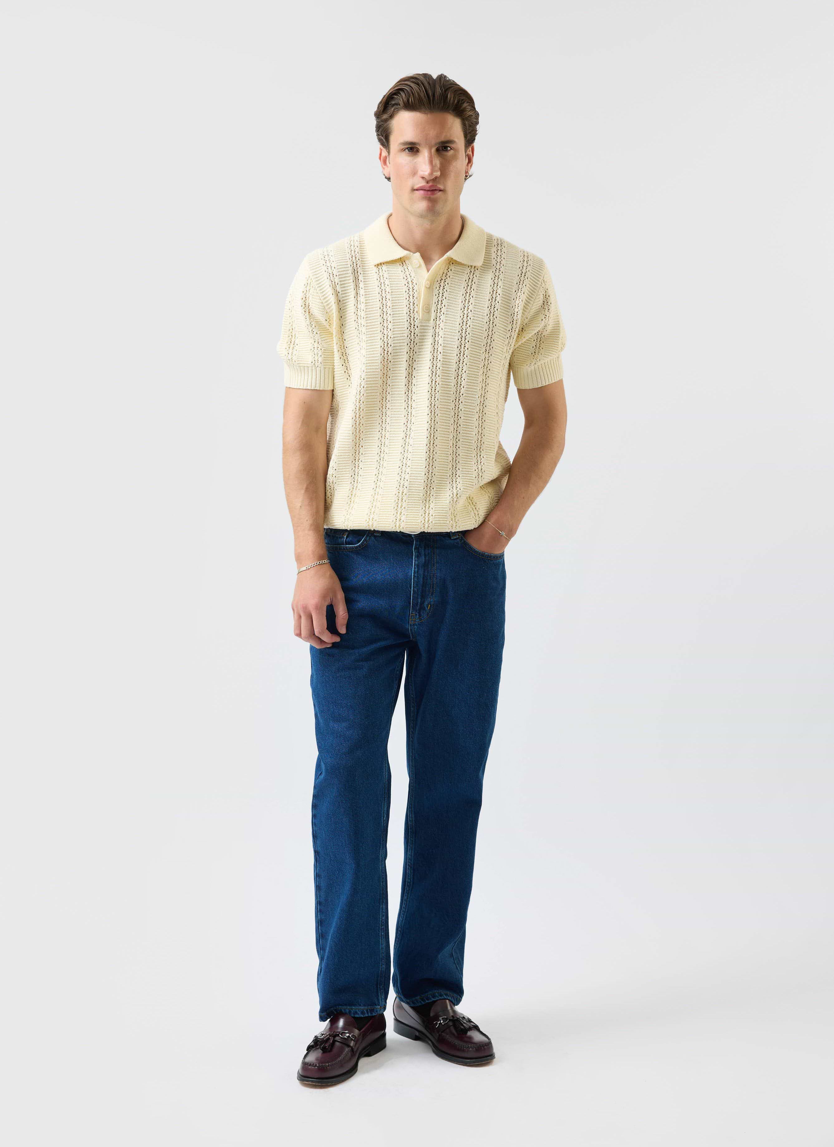 A model wears the Percival Addendum Knitted Polo Shirt in ecru cream, styled with dark blue jeans and loafers.