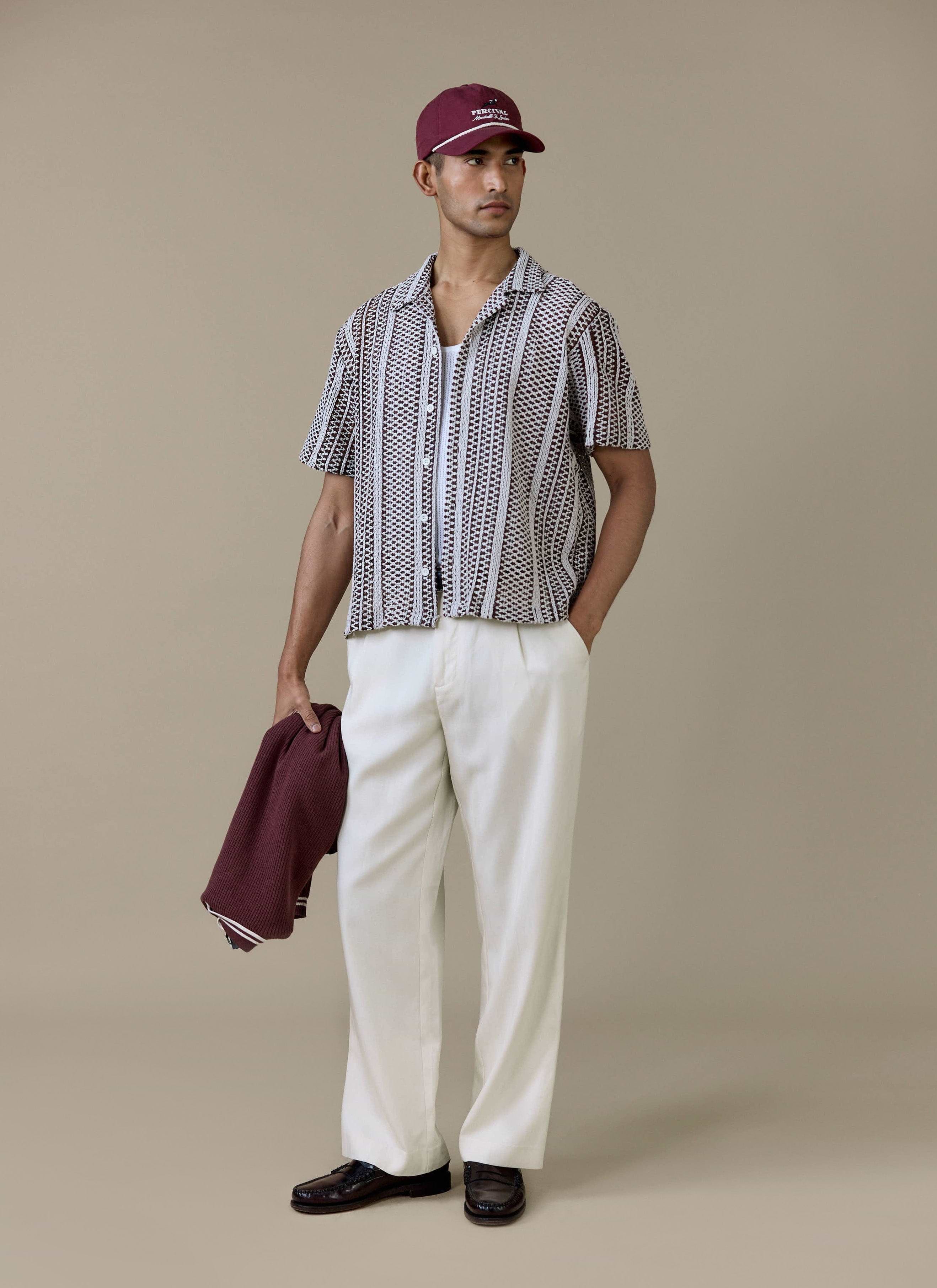 A model wearing the brown Percival Hugh Crochet Boxy Shirt, styled with white trousers and a maroon cap.