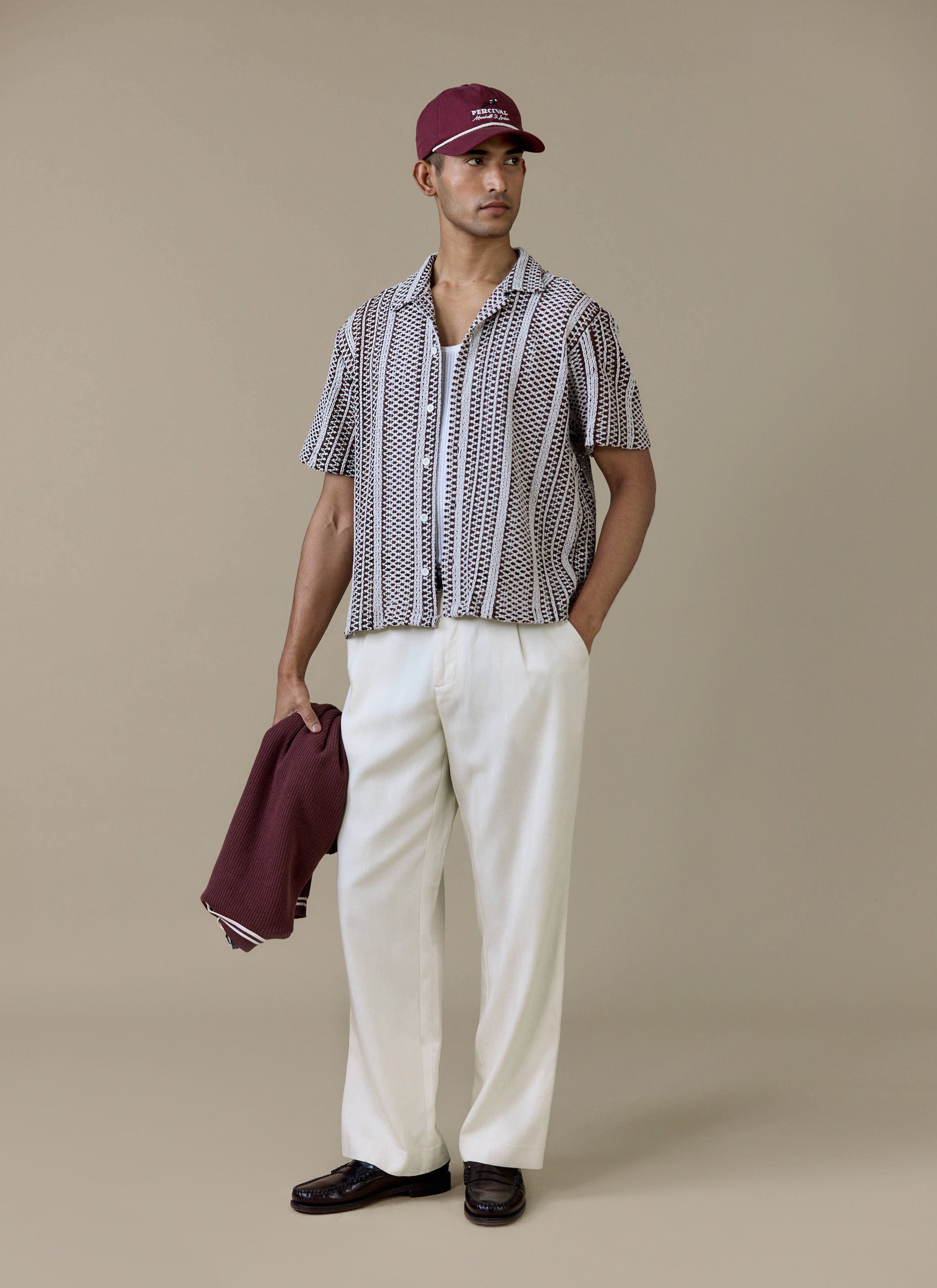 A model wearing the brown Percival Hugh Crochet Boxy Shirt, styled with white trousers and a maroon cap.
