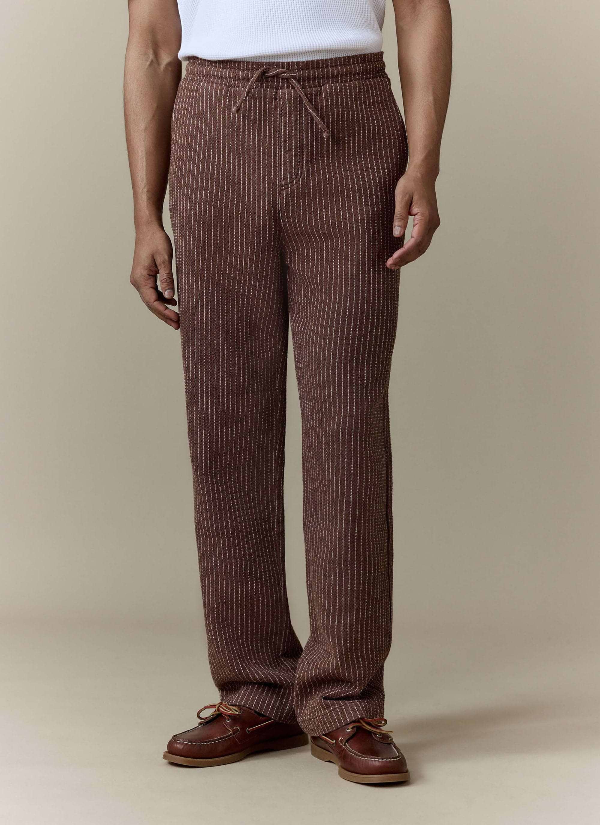 A model wears the brown pinstripe Sorrento Wide Leg Trousers from Percival, styled with a white top and brown boat shoes.