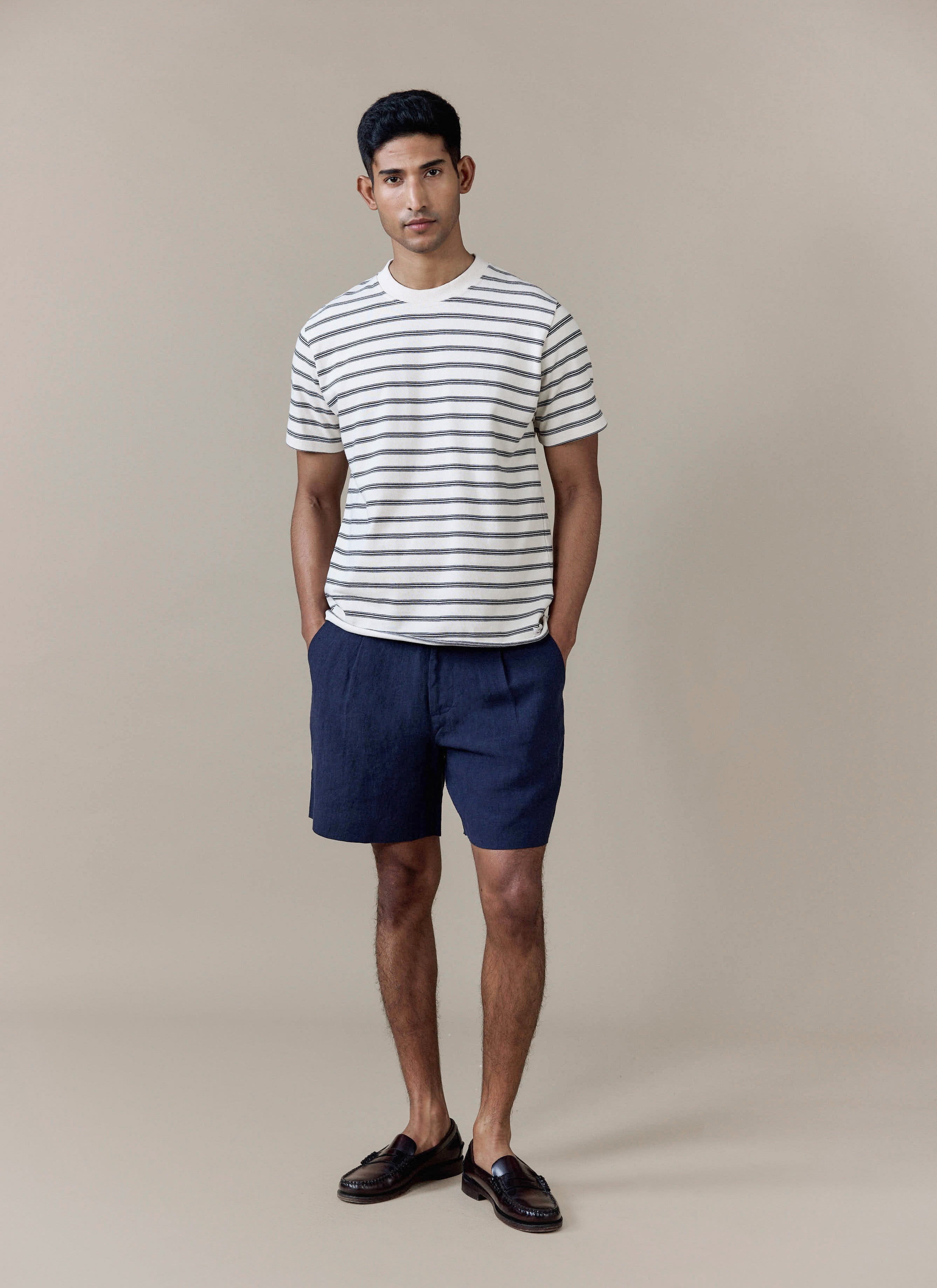 A model wears navy pleated linen shorts by Percival with a striped t-shirt, featuring a tailored fit and front pleats.