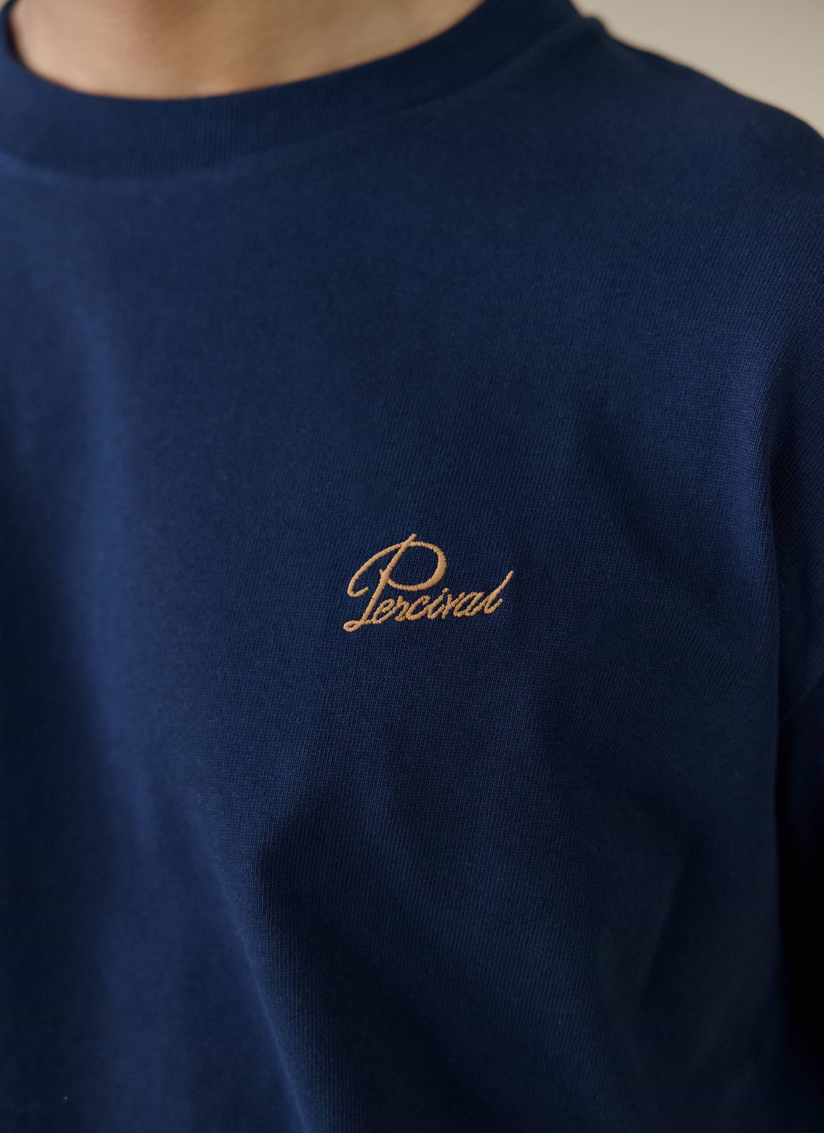 A close-up of the gold script embroidery on the chest of the navy Perci Script T Shirt by Percival.