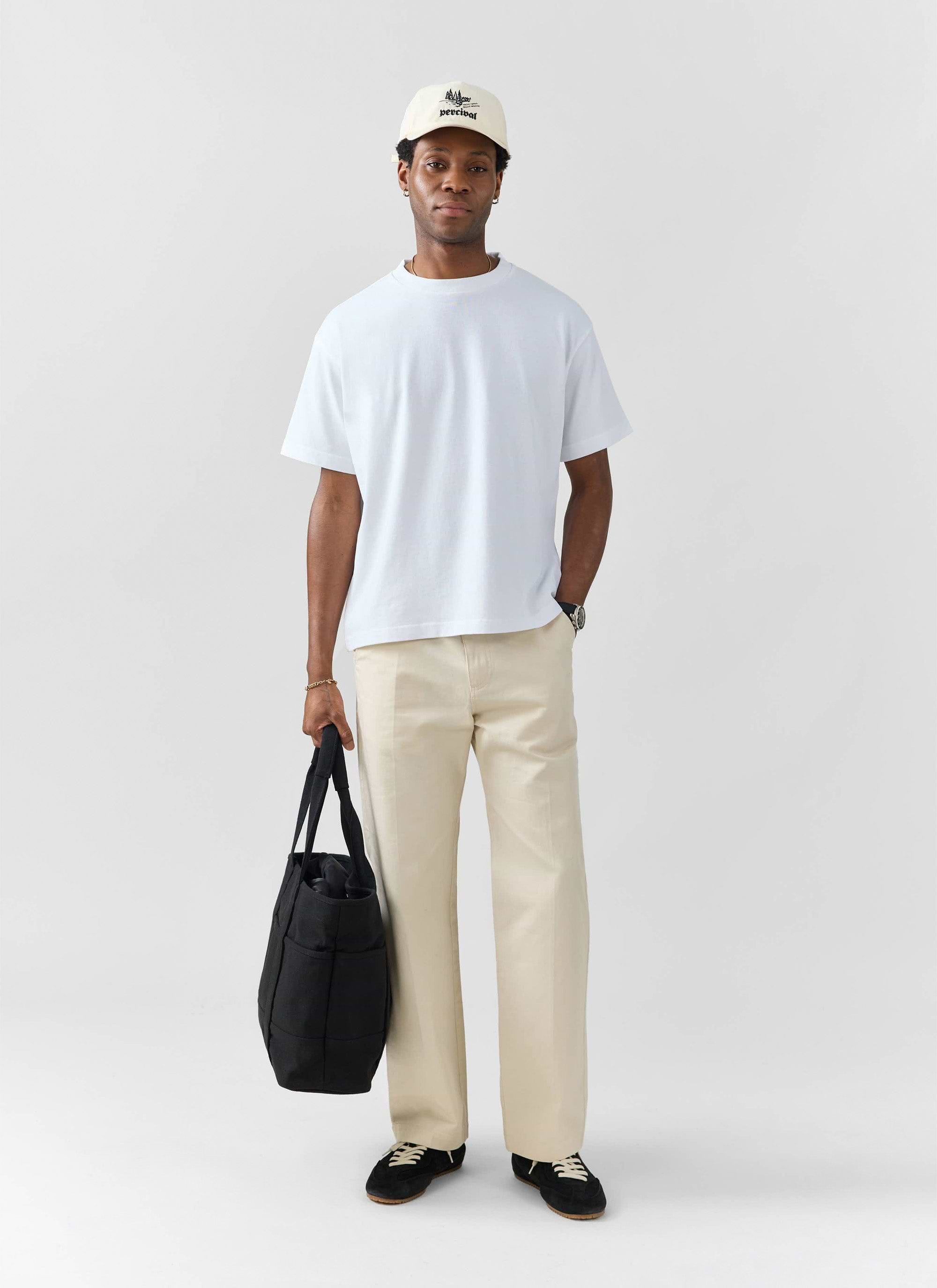 A model wears a white oversized t-shirt from Percival, with cream trousers, black trainers and a cap.