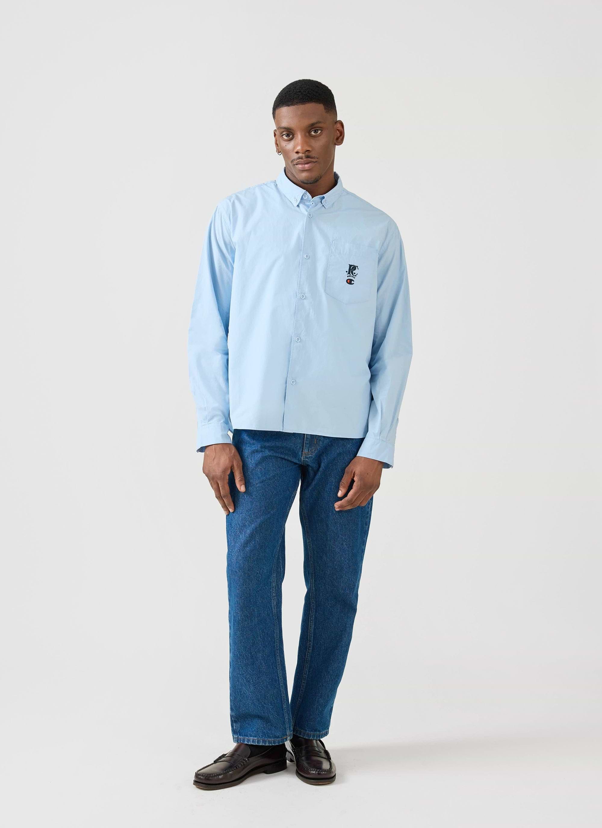 A model wears the light blue Champion x Percival Relaxed Oxford Shirt with blue jeans in a full-length Percival studio shot.