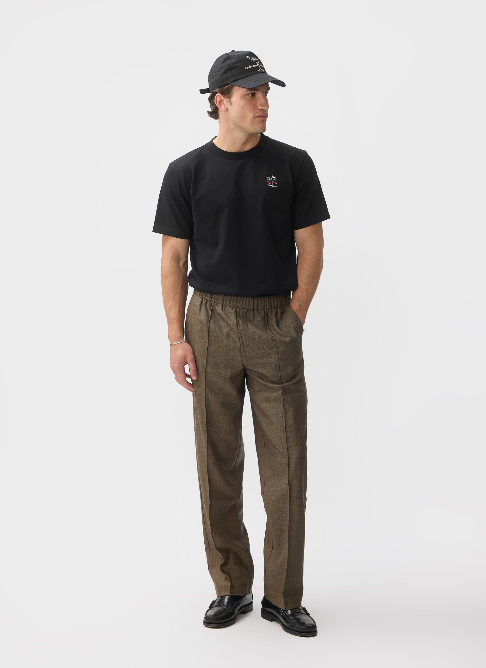 A model wears the black organic cotton À La Carte T Shirt from Percival, styled with brown checked trousers and a black cap.