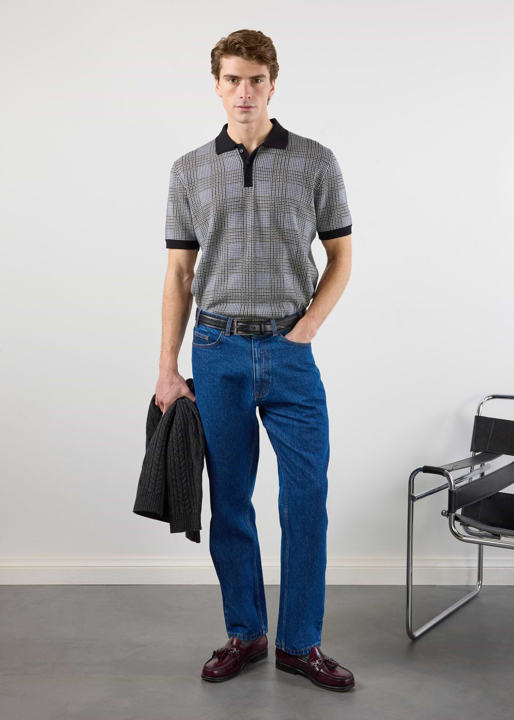 A man models the Percival Casa Martini Knitted Polo in navy puppytooth, styled with blue jeans and loafers in a studio.