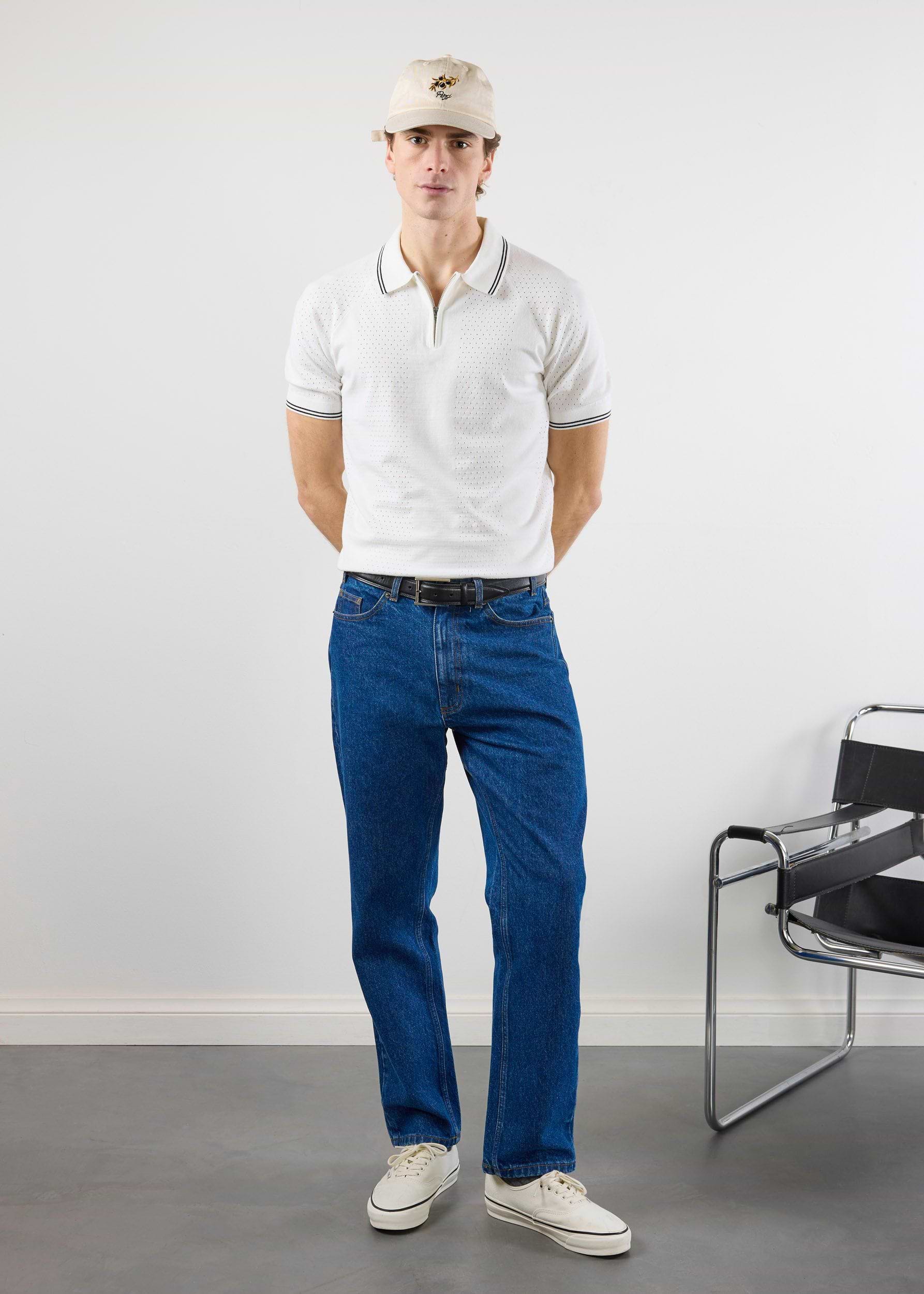 A model wears the Percival Rossi Knitted Polo in white, styled with dark blue jeans, a cap, and white trainers.