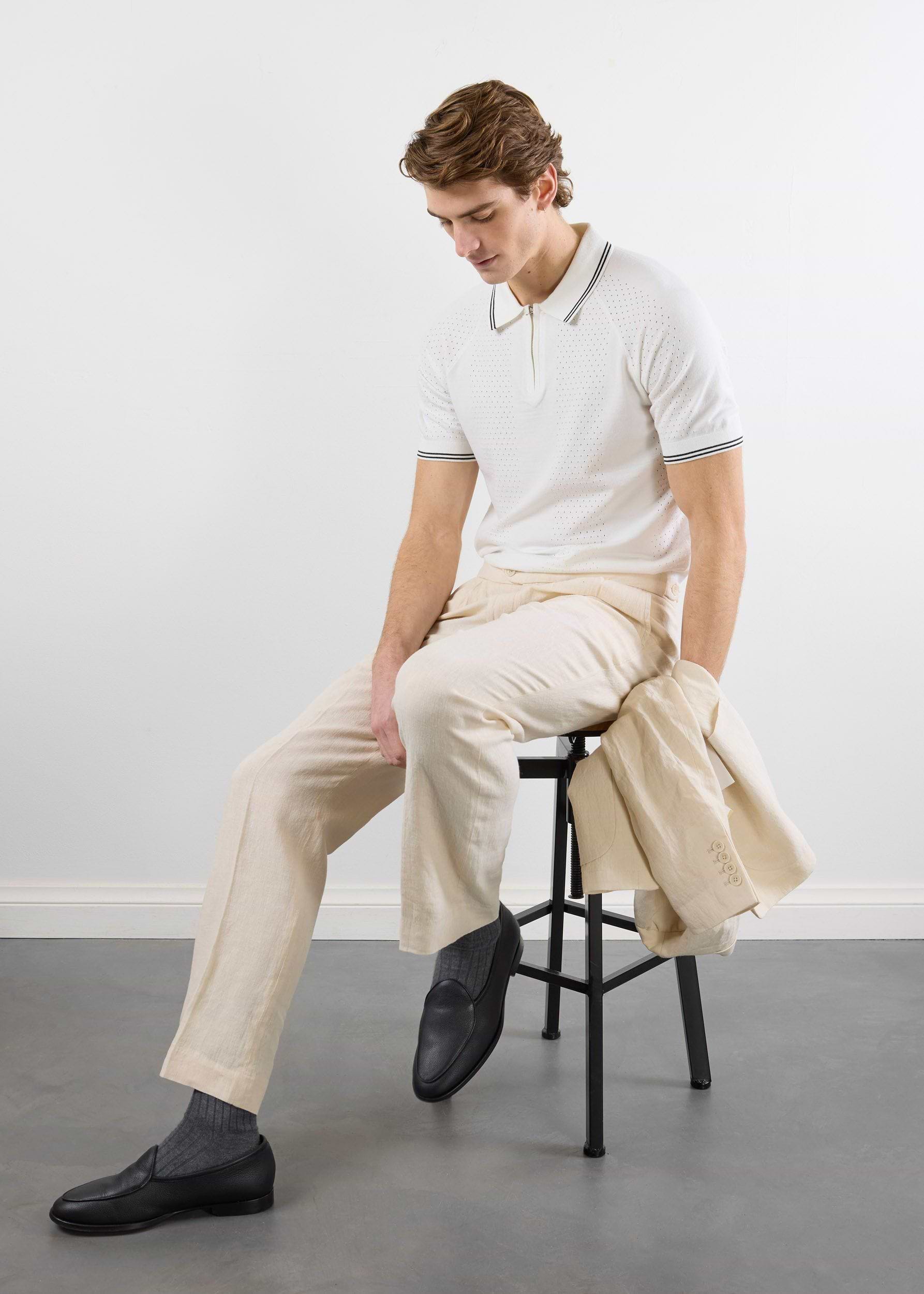 A model sits on a stool wearing the Percival Rossi Knitted Polo in white, paired with ecru trousers and a matching jacket.
