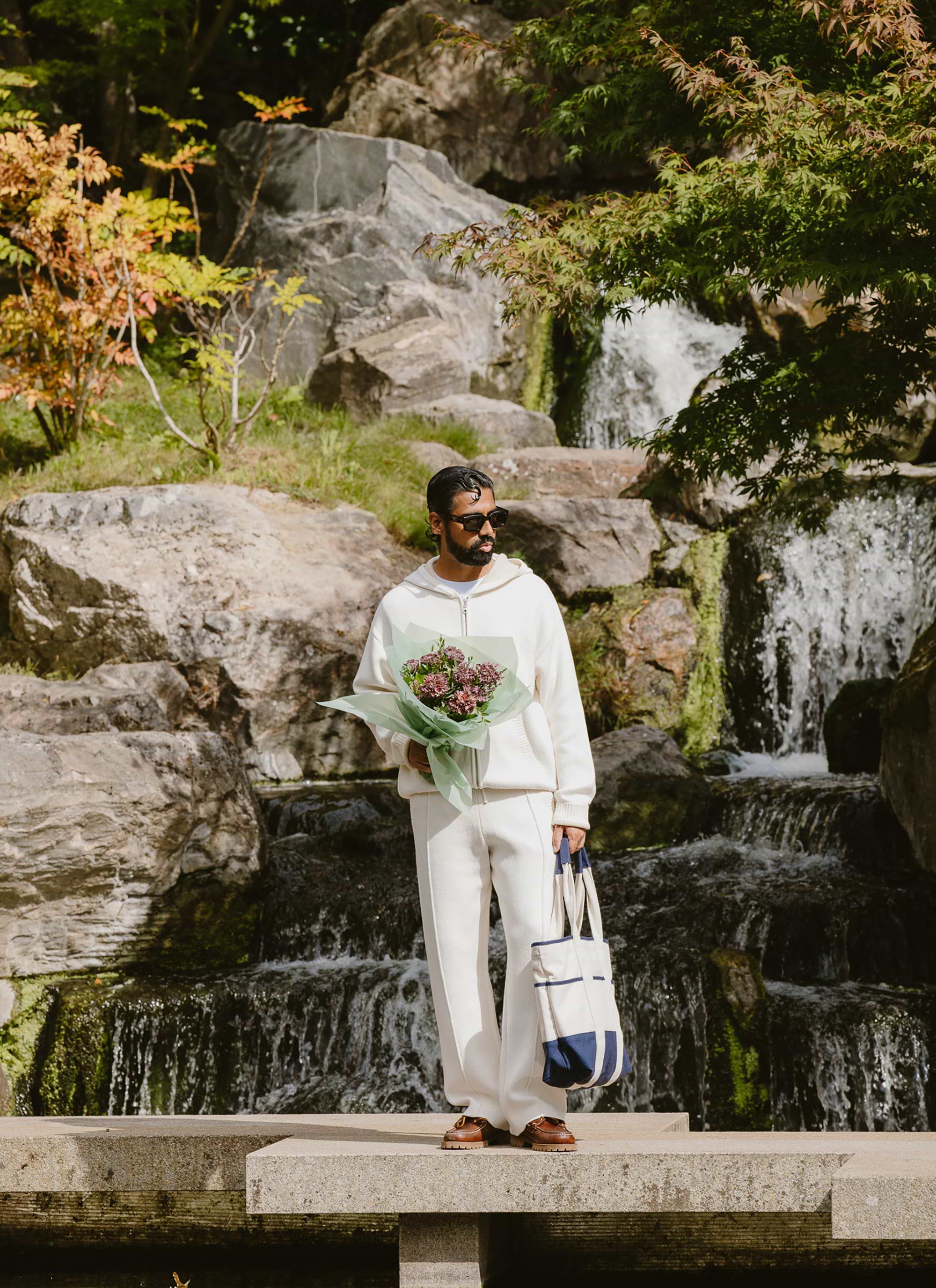 A man models the Percival Chronicle knitted trackpant and hoodie in ecru, standing in front of a waterfall with a bouquet.
