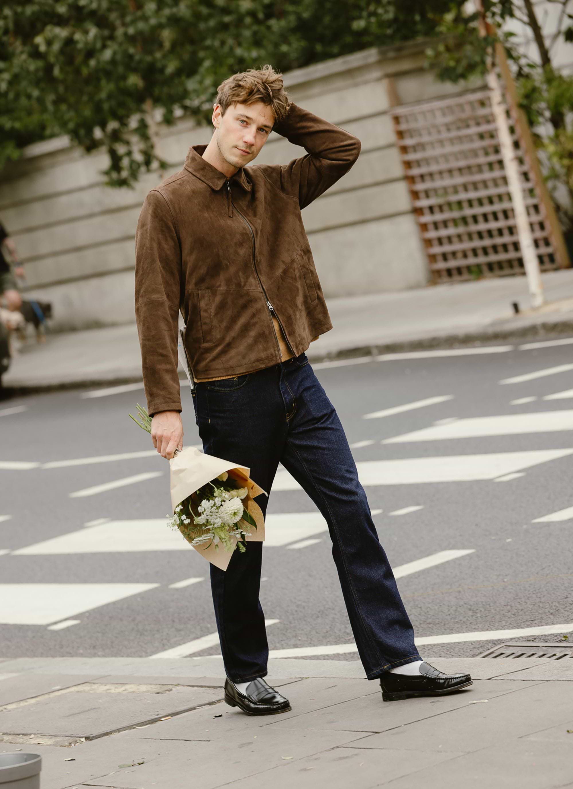 A man models the Percival Prologue Soft Suede Jacket in Espresso Brown on a city street, holding a bouquet of flowers.