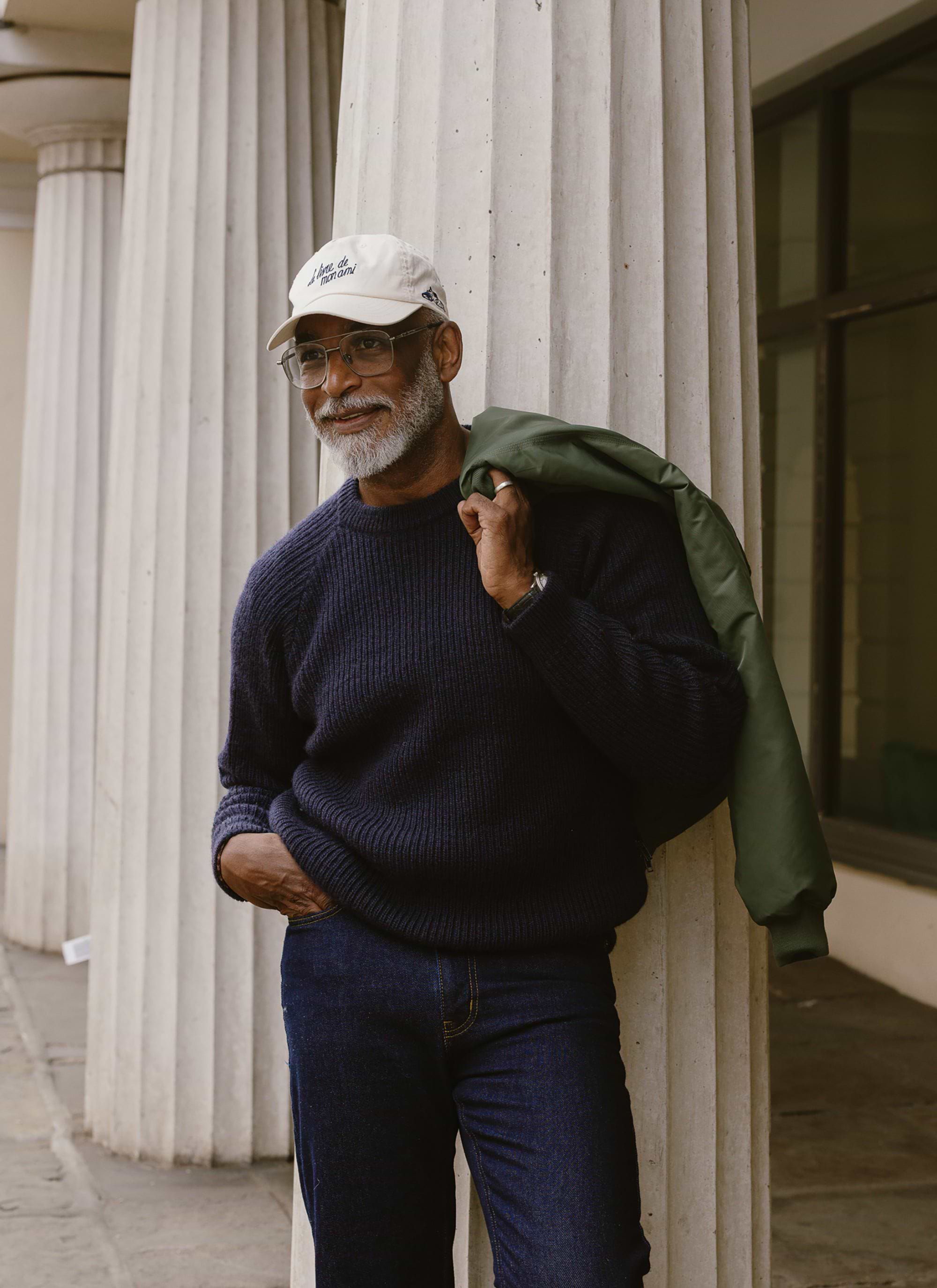A model leans against a pillar wearing the navy wool Fisherman Raglan Jumper from Percival, with a jacket over his shoulder.