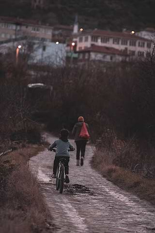 A child rides his bike on the road, following his mother who is walking.