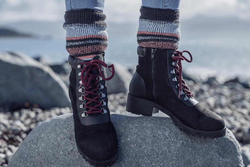 Close-up of a person's legs standing on a rock, wearing black boots with red laces, and thick, striped Heat Holders ®thermal socks