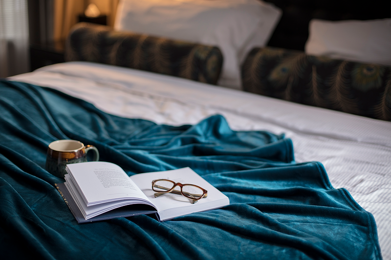 Glasses on a book with a coffee cup on a rich thermal blanket in a bedroom.