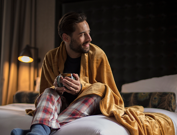 Man wrapped in fleece blanket, siting on a bed, holds mug.