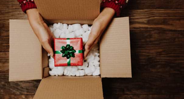 A woman's hands removing a Christmas gift surrounded by foam peanuts in a plain box.