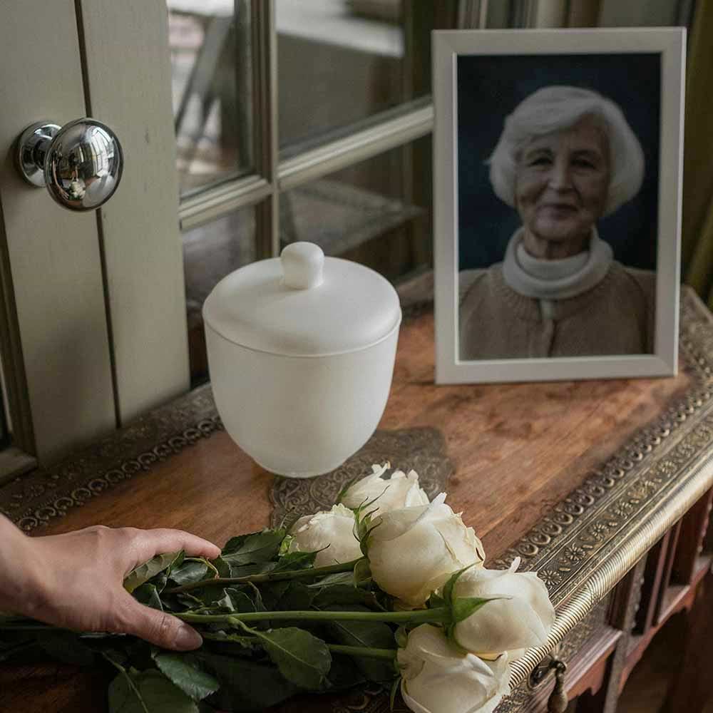 Urn with ashes on a table, a memorial photo, and a hand holding flowers.