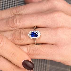 Vintage 1920 style engagement ring with diamond halo and bezel set 2ct oval cut lab created blue sapphire in 14k white gold worn on hand