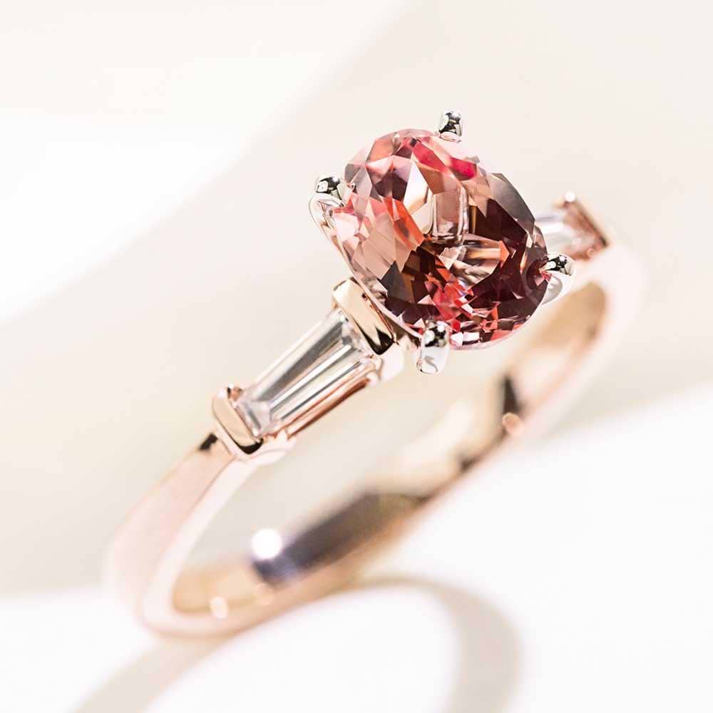 Shown with 1.5ct Oval Cut Lab Grown Champagne Pink Sapphire in 14k Rose Gold|Three stone engagement ring with 1.5ct oval cut lab grown pink sapphire amid two baguette cut diamond side stones in 14k rose gold