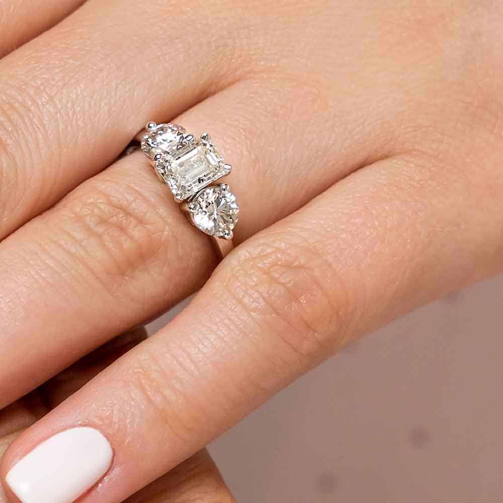 Shown with 2.5ct Emerald Cut Lab Grown Diamond Center Stone in 14k White Gold|Three stone engagement ring with 2.5ct emerald cut center stone surrounded by round cut lab grown diamonds on either side worn on hand